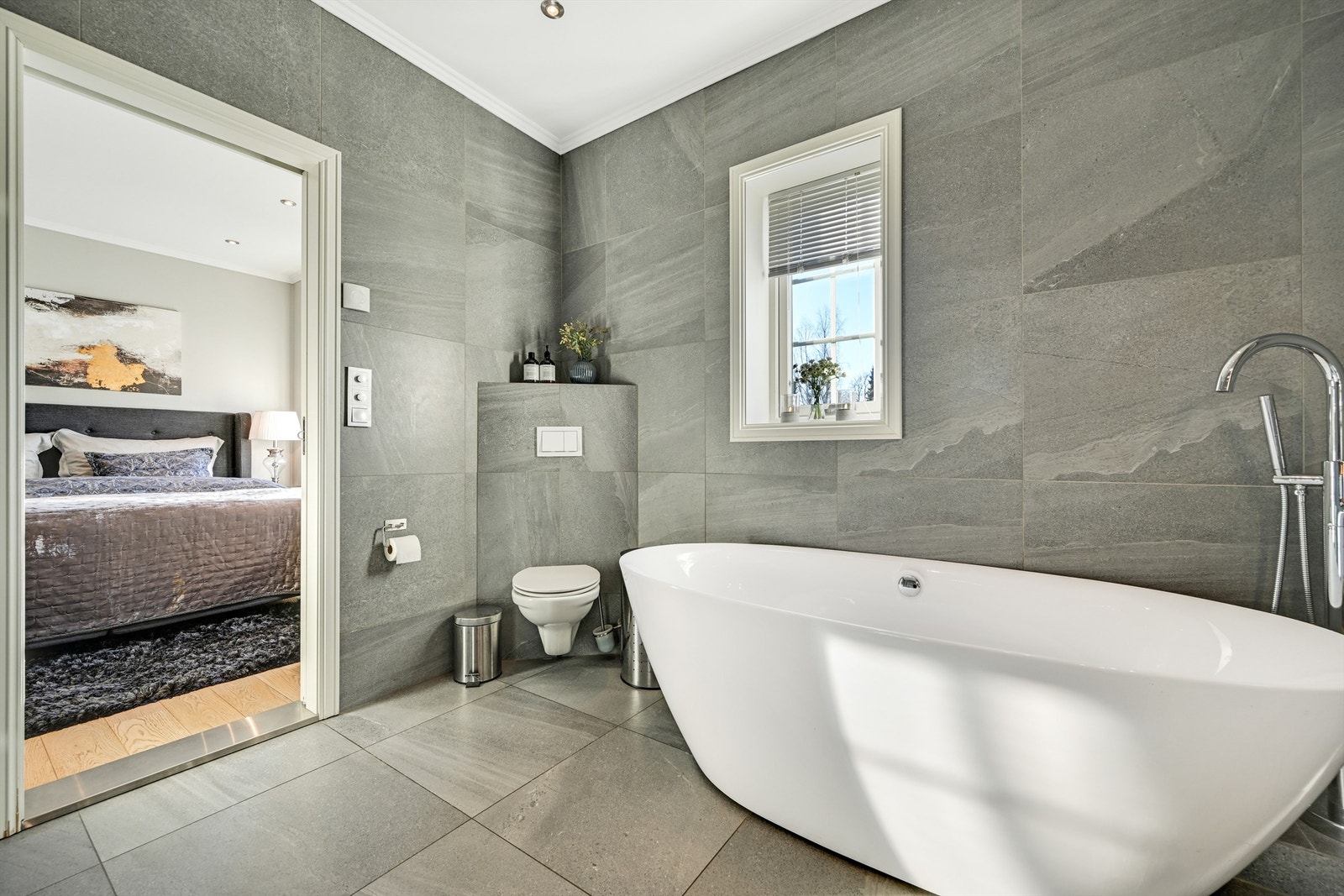 Badet er innredet med fliser på gulv og vegger, og har dobbel servant, veggmontert toalett, badekar samt dusjhjørne med glassvegger. Galleribilde