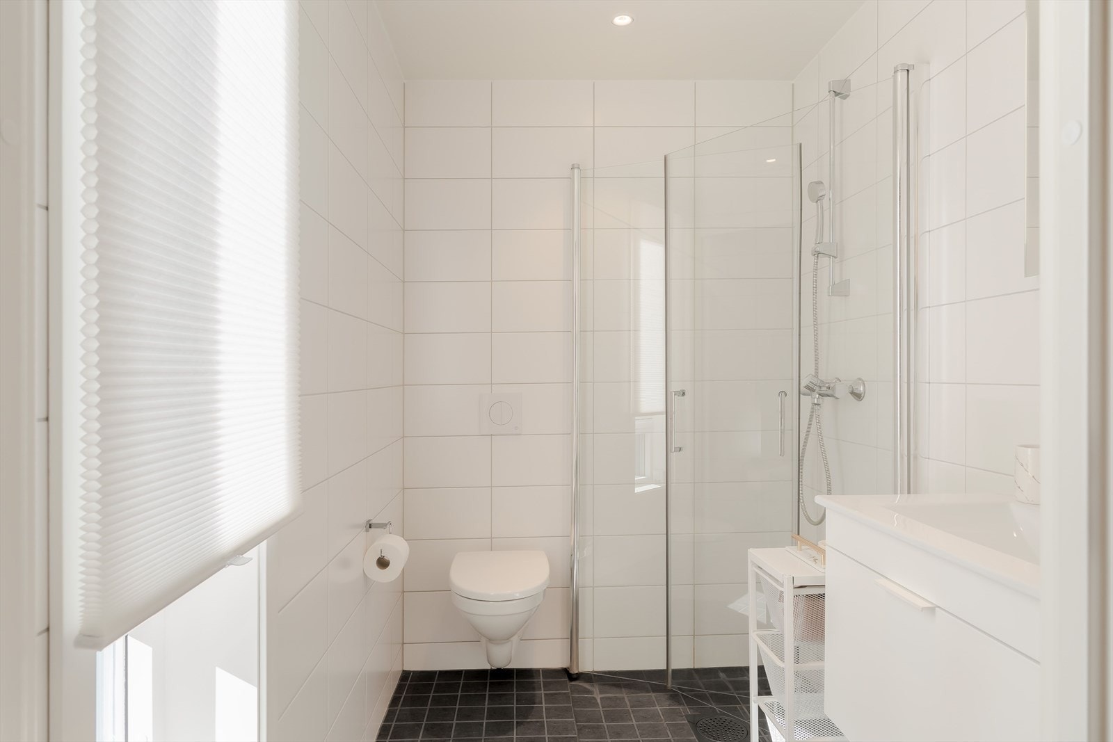 Også dette badet har et helhetlig uttrykk med hvite veggfliser, mørkt flisgulv og elektriske varmekabler, samt vegghengt toalett, servantinnredning og dusjsone med glassvegger. Galleribilde