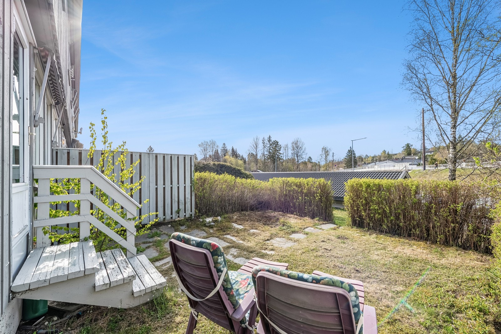 Utgang fra stuen med trapp til hellelagt solplass og overgang til hage skjermet med hekk og beplantet med prydbusker. Galleribilde