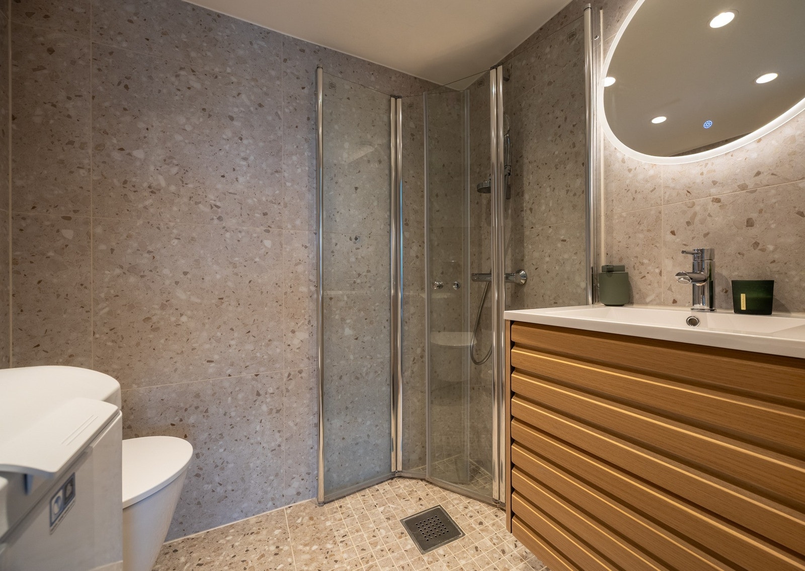 Delikat bad med steinmønstrede fliser på gulv og vegger. Badet ha downlights, varmekabler, samt et dusjhjørne med mosaikkfliser på gulv og foldedører i glass. Galleribilde