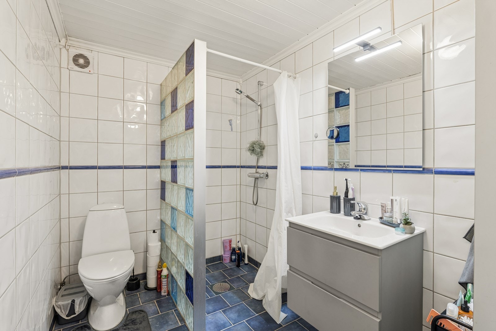 Innredningen består av nedfelt servant, toalett og opplegg for vaskemaskin og tørketrommel. Det er glassbyggerstein og forheng inn til dusjsonen. Galleribilde