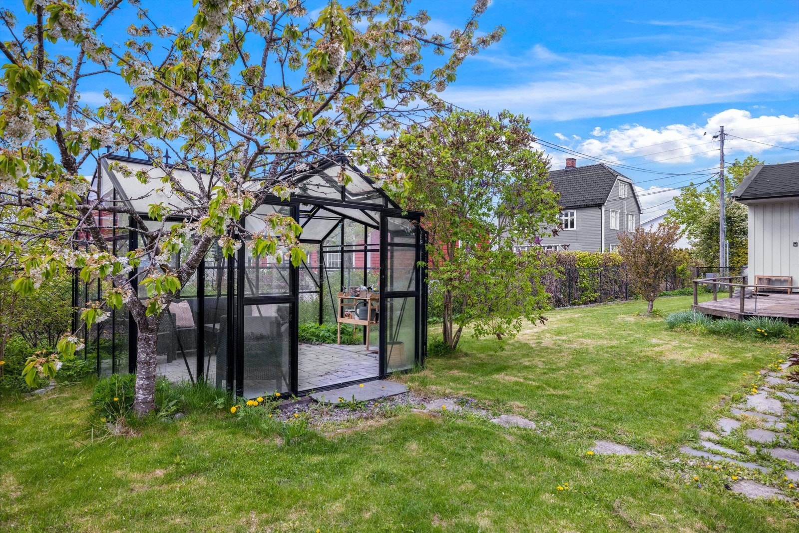 Flat, opparbeidet tomt rundt huset med plen, busker, blomster og trær. Gangsti med steinheller og romslig drivhus. Galleribilde