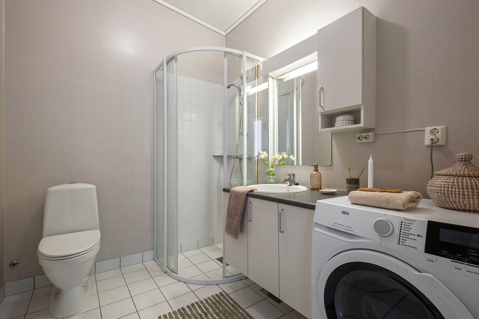 Badet har fliser og elektriske varmekabler. Det er tapetserte veggplater på vegger med flis i dusjsonen og malt innvendig tak. Rommet er innredet med servantinnredning, toalett og dusjhjørne med glassdør. Opplegg for vaskemaskin. Det er mekanisk avtrekk. Galleribilde