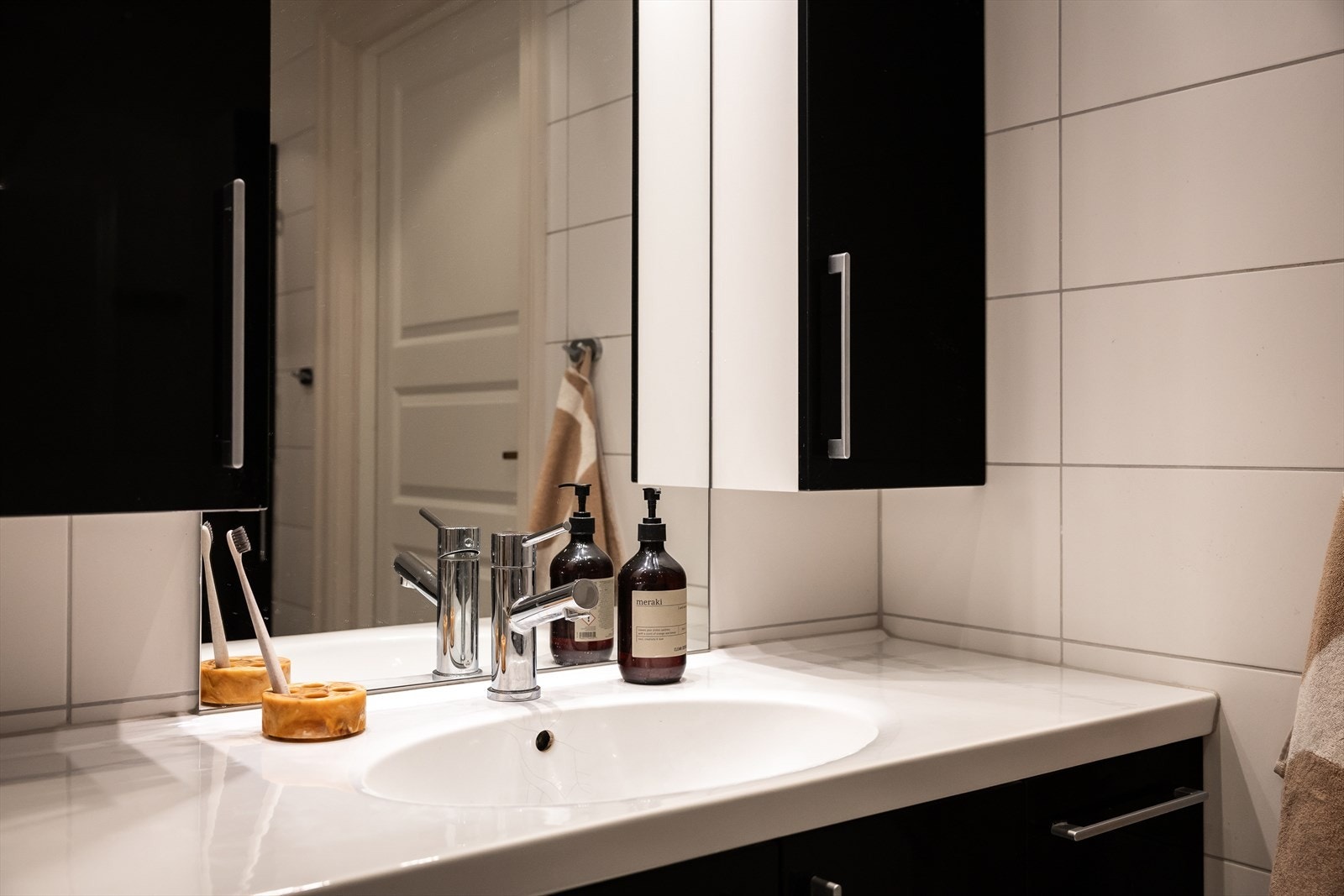 Detalj fra badet: Servant med heldekkende vask og krom armatur, vegghengt skap i kontrastfarger og store, lyse fliser gir et rent og moderne uttrykk. Galleribilde