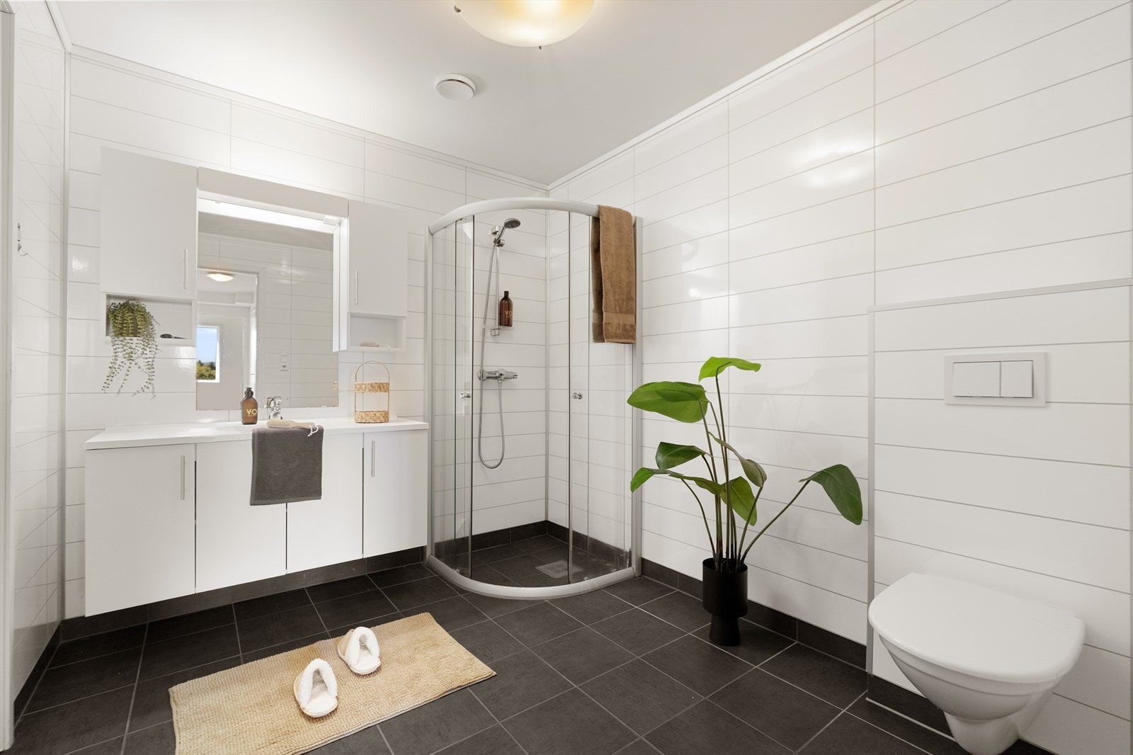 Badet inneholder baderomsmøbel med slette fronter, heldekkende servant, speilskap med belysning, vegghengt toalett og buet dusjhjørne med dusjgarnityr. Galleribilde