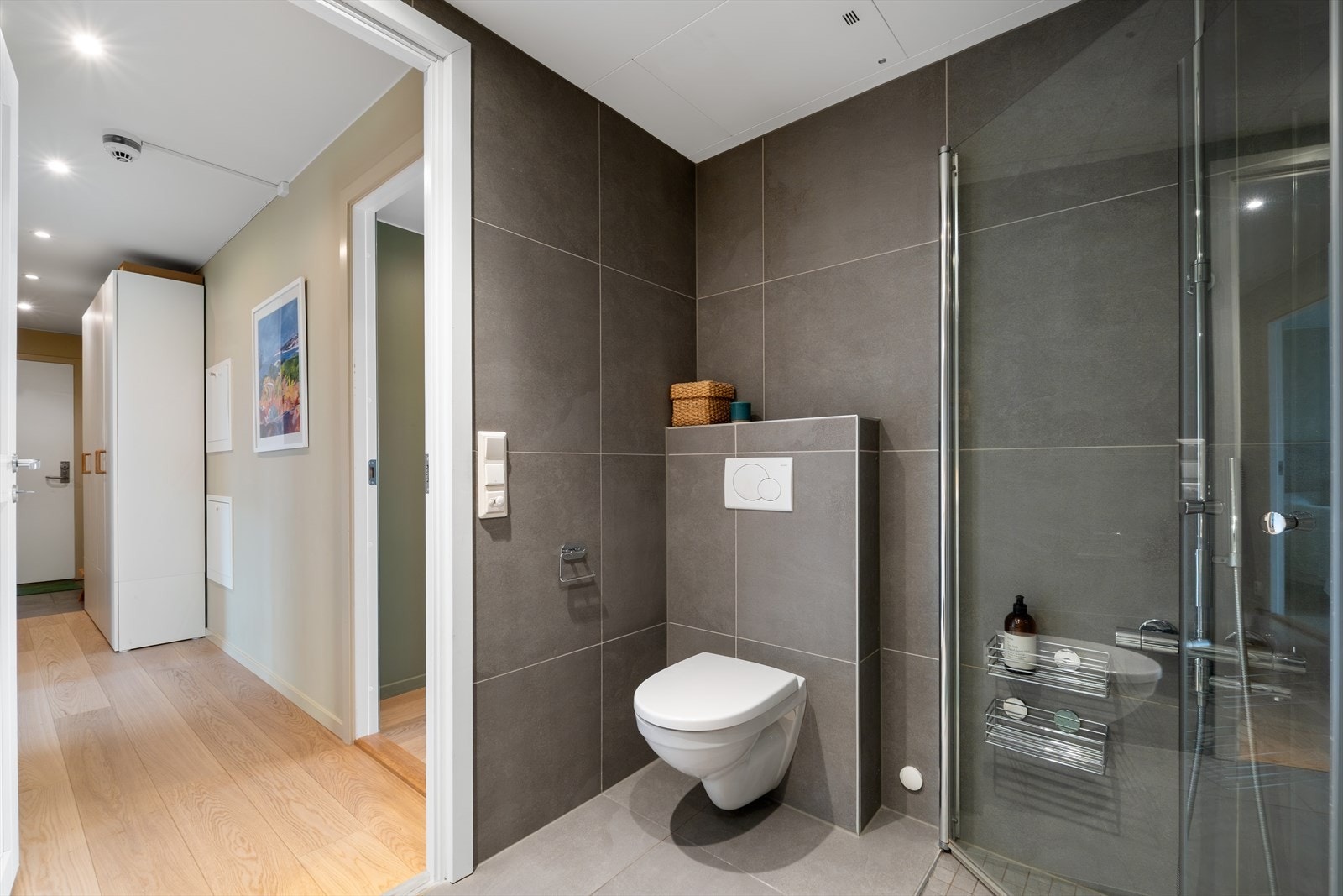 Videre har badet veggmontert wc, opplegg for vaskemaskin og et dusjhjørne med innfellbare glassdører og hånd- og regndusjarmatur. Badet har mekanisk avtrekk. Galleribilde