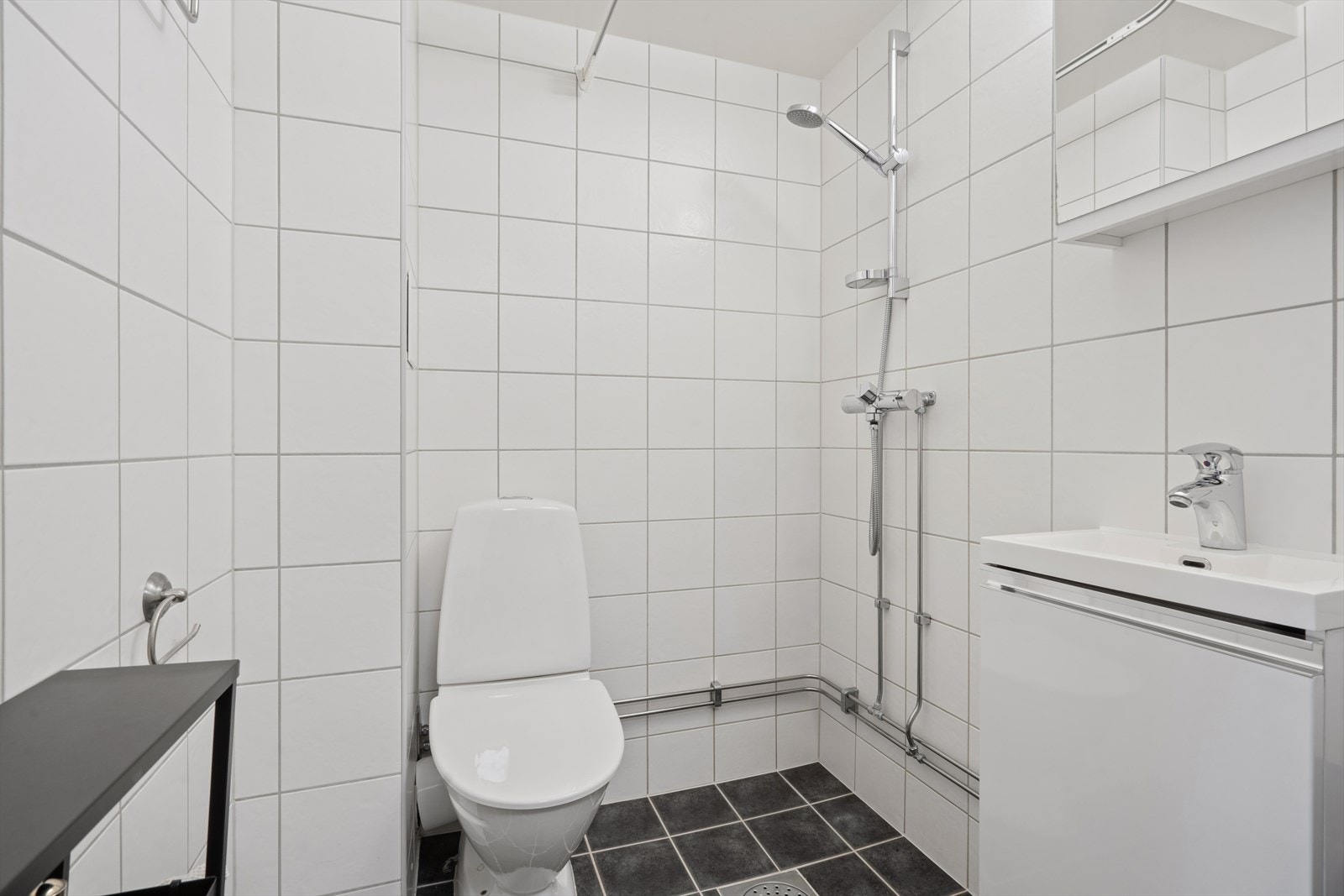 Badet er innredet med gulvstående toalett, dusj direkte på gulv med forheng, samt servant med underskap og tilhørende kran. Galleribilde