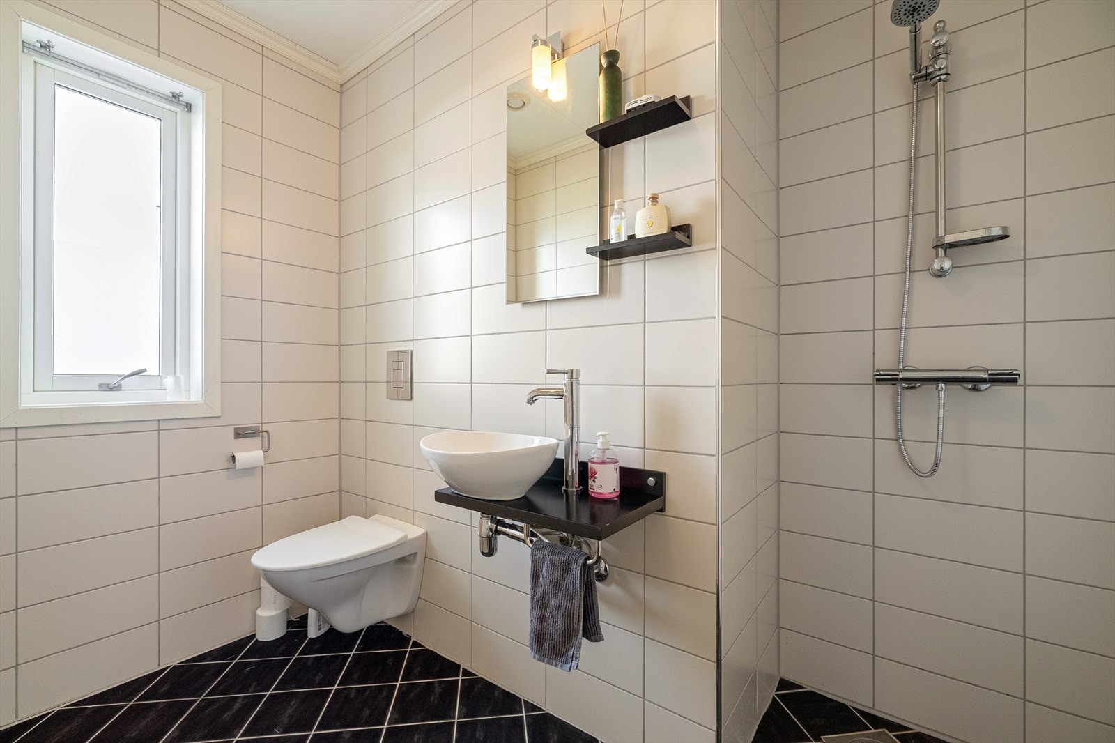 Baderommet er helfliset og inneholder innredning med benkeplate i laminat, toppmontert servant, speil med vegglampe, vegghengt toalett og dusj. Galleribilde