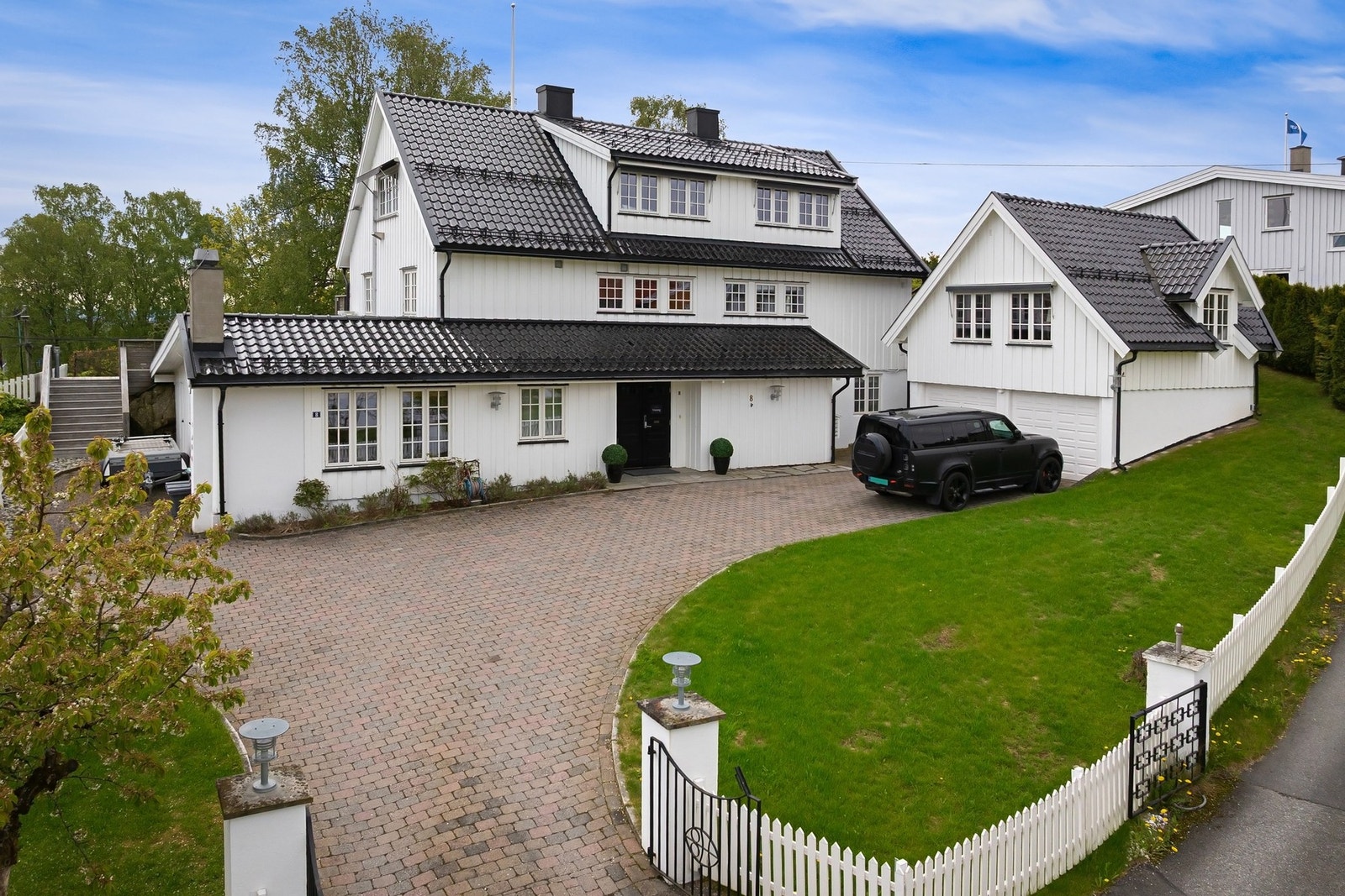 Bred, steinsatt gårdsplass bak smijernsport og hvitmalt stakitt - et ankomstparti som gir både et representativt førsteinntrykk og en følelse av privatliv. Galleribilde