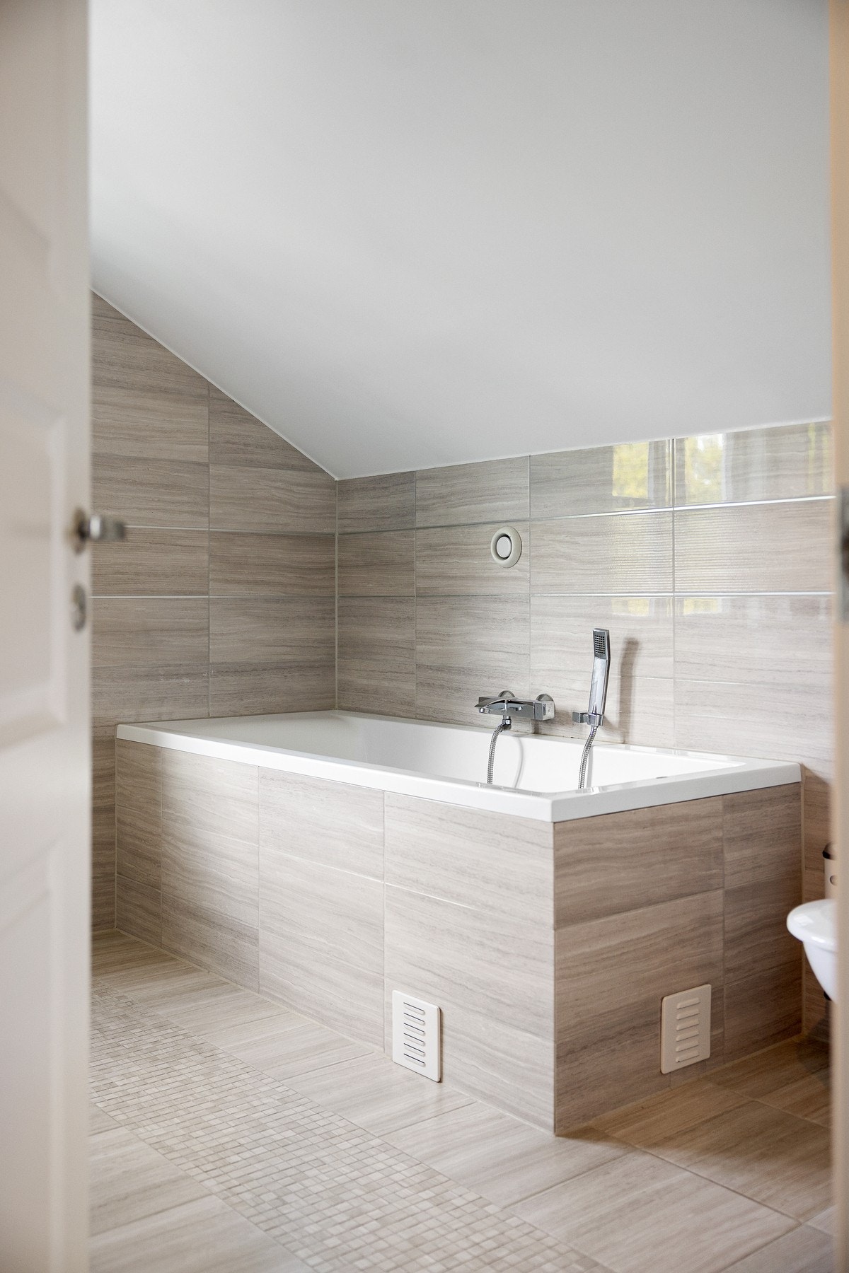 Badet har gulvvarme, downlights og naturlig dagslys fra vindu. Innredet med innmurt badekar, vegghengt toalett og moderne servantinnredning med hvite skuffer og speil med integrert belysning. Galleribilde