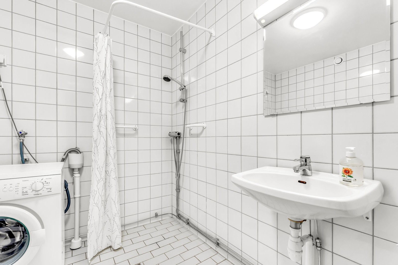 Lyst bad med dusj, vaskemaskin og servant. Flislagte vegger og gulv. Galleribilde