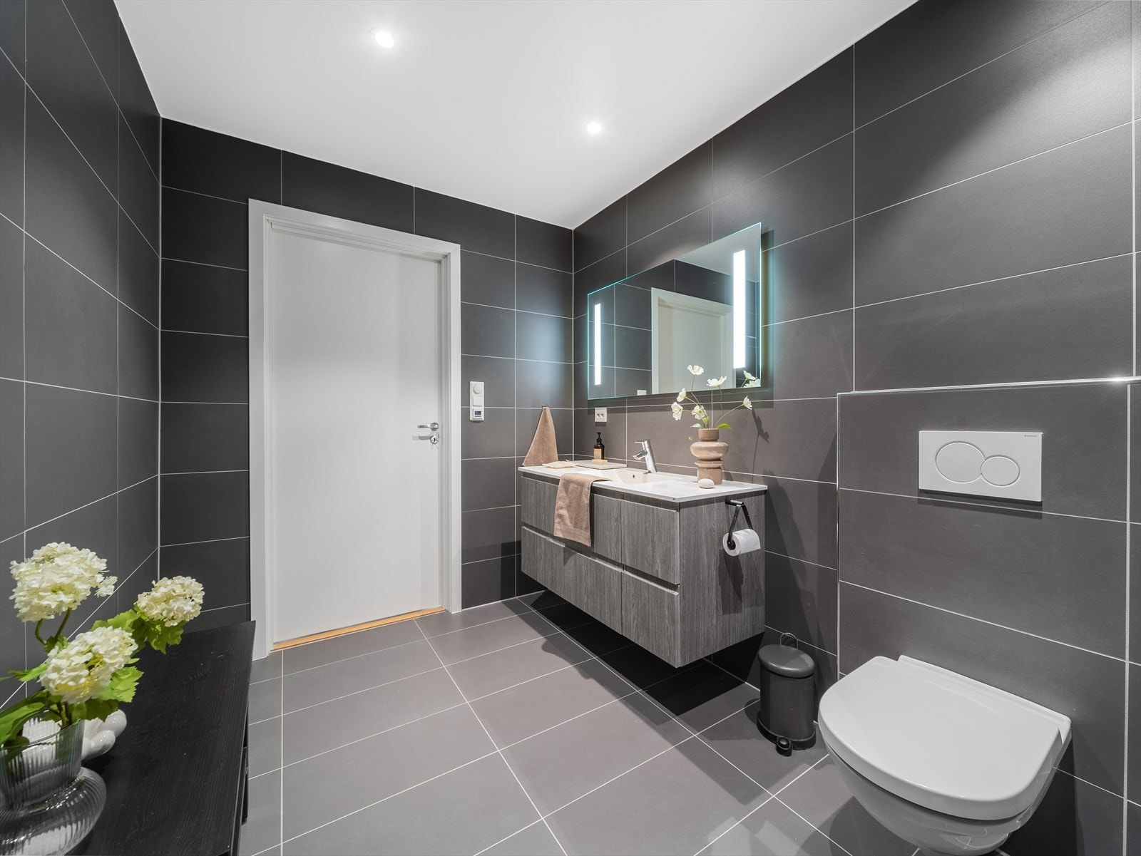 Badet er utstyrt med dusjvegg i glass til nedsenket dusjsone, vegghengt toalett med innebygd sisterne og moderne servantmøblement med speil og integrert belysning. Galleribilde