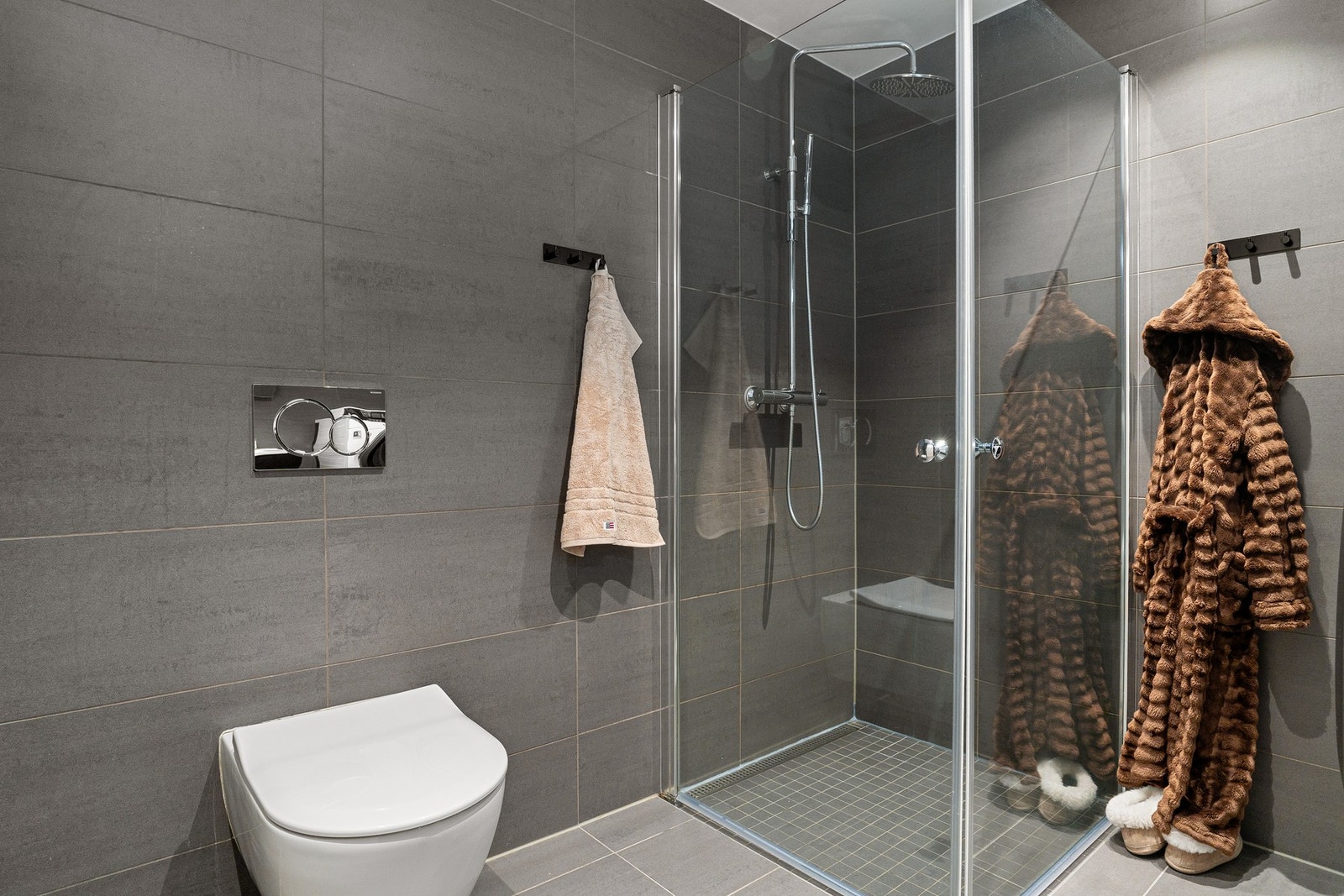 Badet ved soverom har tilsvarende flislagt og gjennomført uttrykk med servantskap med heldekkende servant, speilskap med lysarmatur, veggmontert toalett og dusjhjørne med svingdører, regnfallsdusj og termostatstyrt blandebatteri, samt gulvvarme. Galleribilde