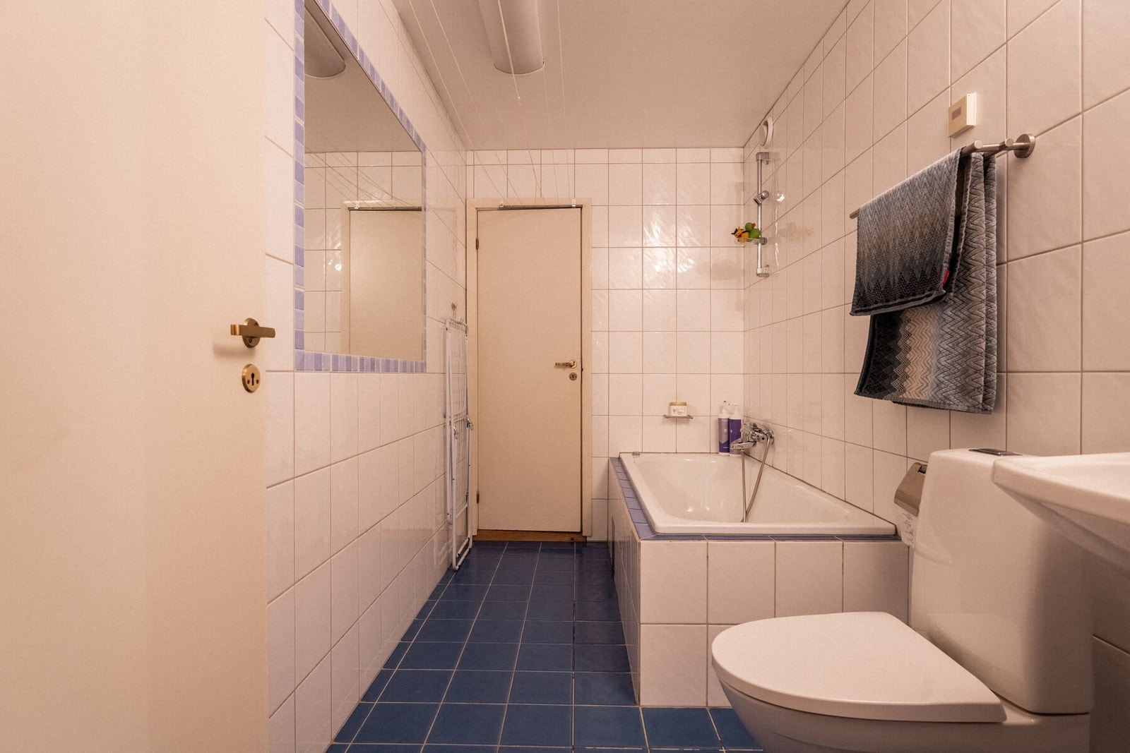 Flislagt bad fra byggeår med innbygget badekar, gulvmontert toalett (skiftet 2023) og servant med speil. Galleribilde