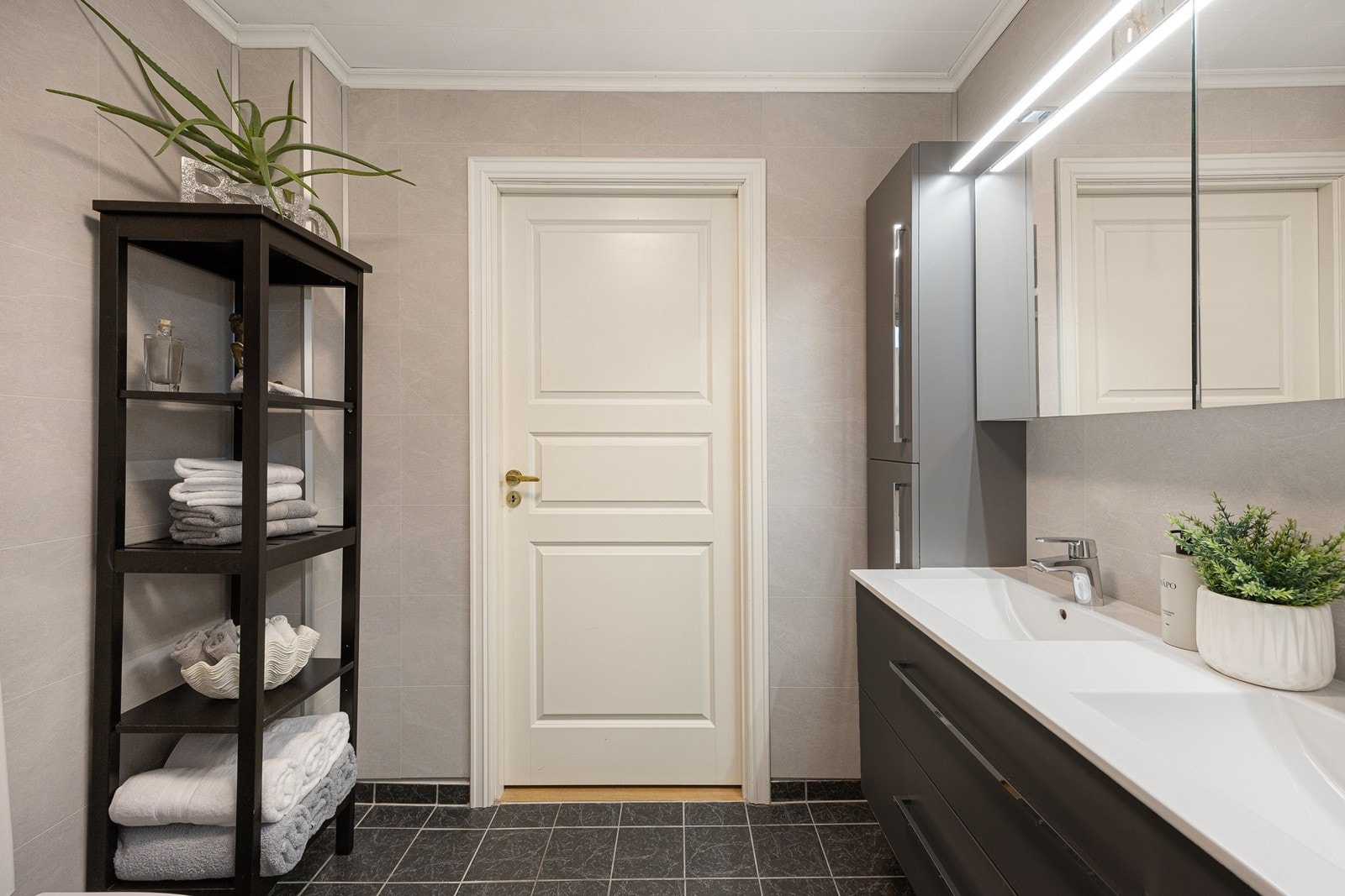Videre har badet et toalett, et dusjkabinett og opplegg for vaskesøyle. Badet fremstår som velholdt og pent, med lyse våtromsplater på veggene og grå fliser på gulv med gulvvarme. Galleribilde