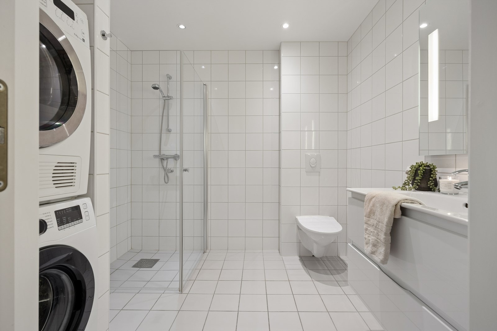 Moderne og tidløst, helfliset bad/vaskerom med vegghengt toalett, dusjhjørne, baderomsinnredning og opplegg for vaskemaskin. Galleribilde