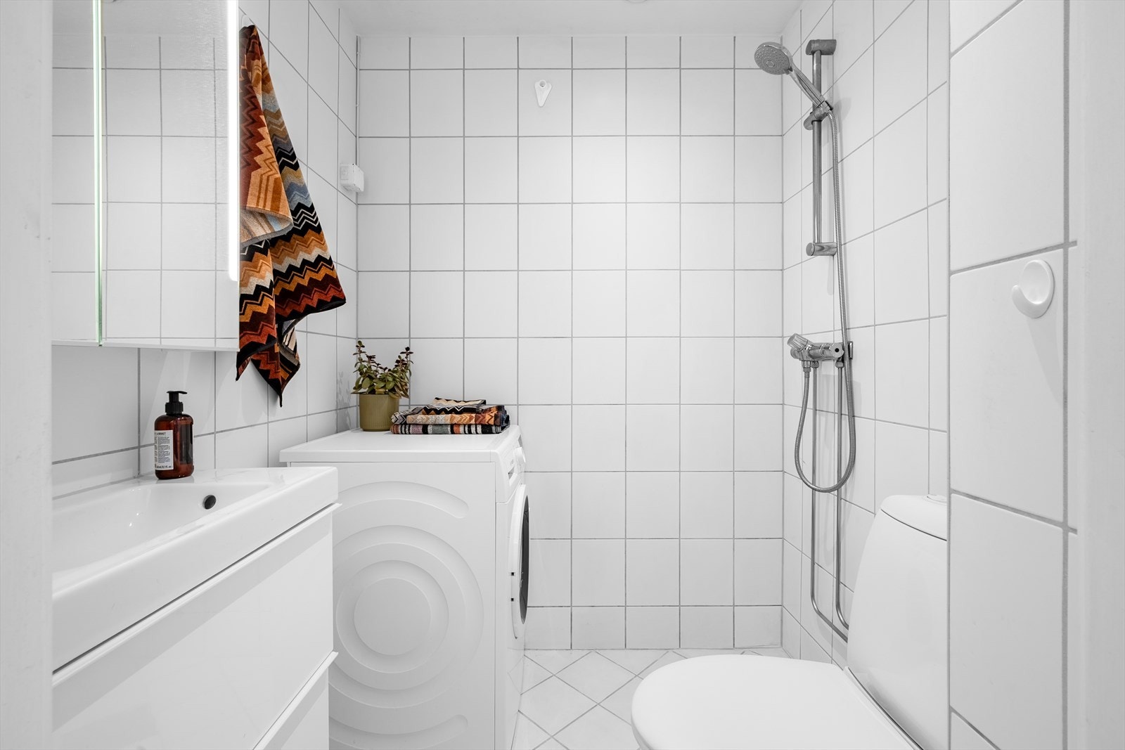 Et lyst og flislagt baderom med downlights i himling og opplegg for vaskemaskin. Galleribilde