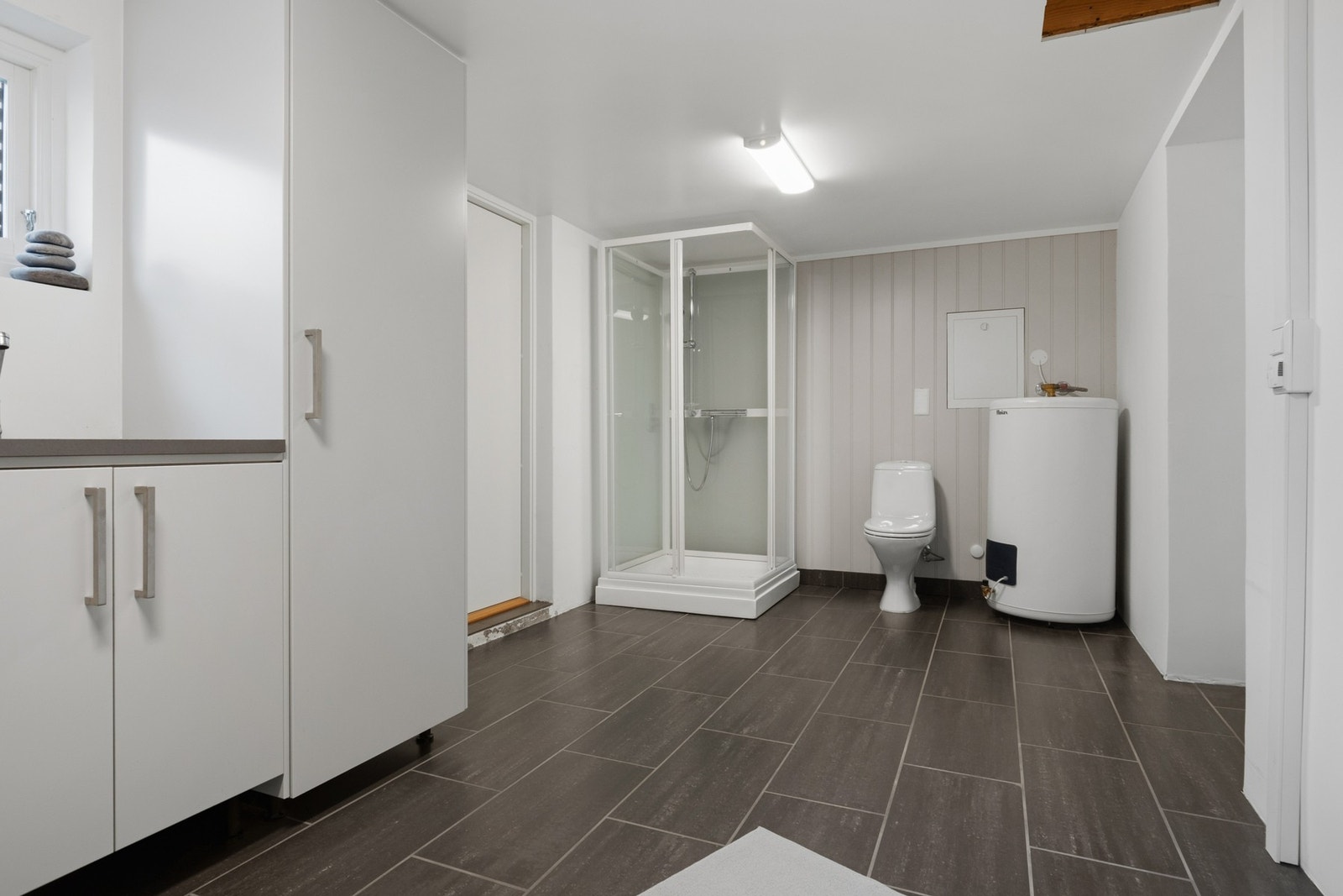 Praktisk kombinert vaskerom og bad med flislagt gulv, varmekabler, dusjkabinett, toalett og opplegg for vaskemaskin. Galleribilde