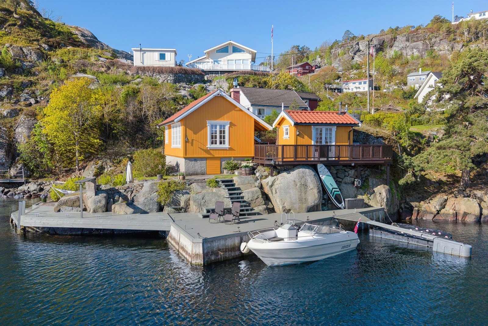 Eiendommen sett fra sjøsiden, med hytte, uthus og et stort bryggeanlegg. Galleribilde