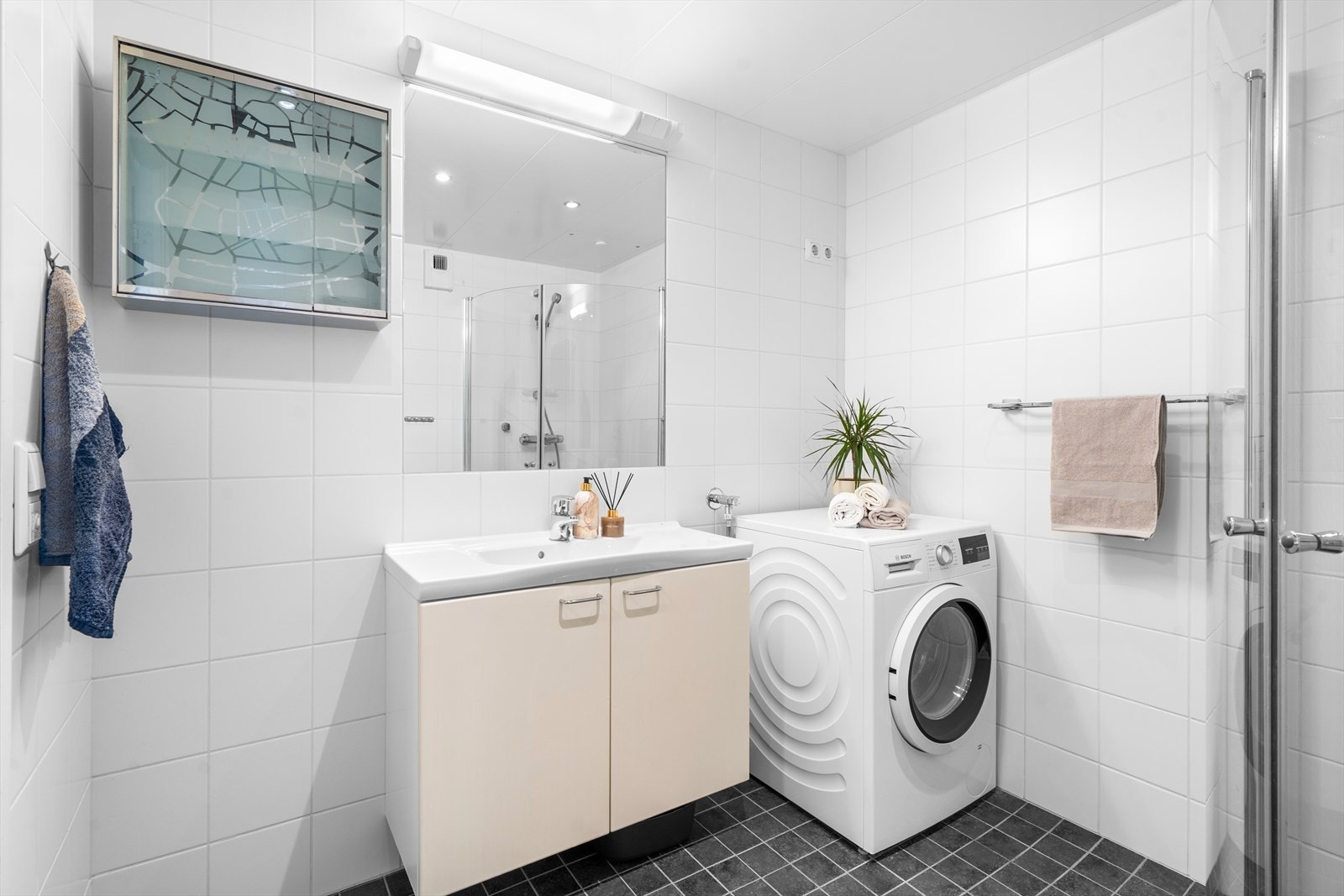 Badet er innredet med veggmontert toalett, dusj, speil med overliggende belysning, baderomsinnredning med malte glatte fronter og heldekkende servant. Galleribilde