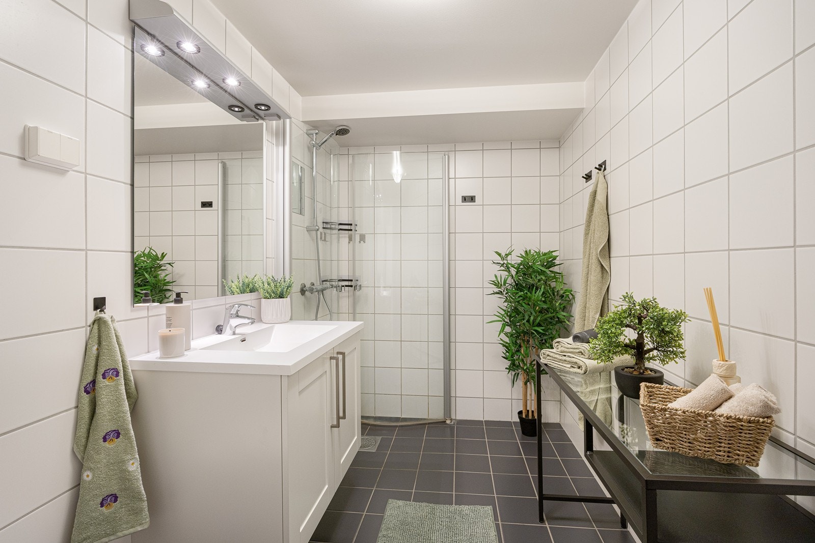 Baderomsinnredningen består av heldekkende servant med hvite underskap og et speil med tilhørende lys. Badet har vegghengt toalett og et dusjhørne med innfellbare glassdører. Galleribilde