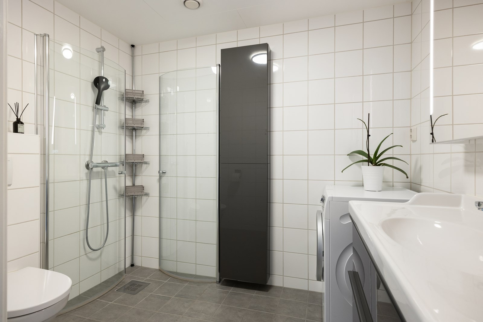Badet er innredet med veggmontert toalett, dusjhjørne med glassdører, høyskap, servant med baderomsinnredning og speil med integrert belysning. Galleribilde