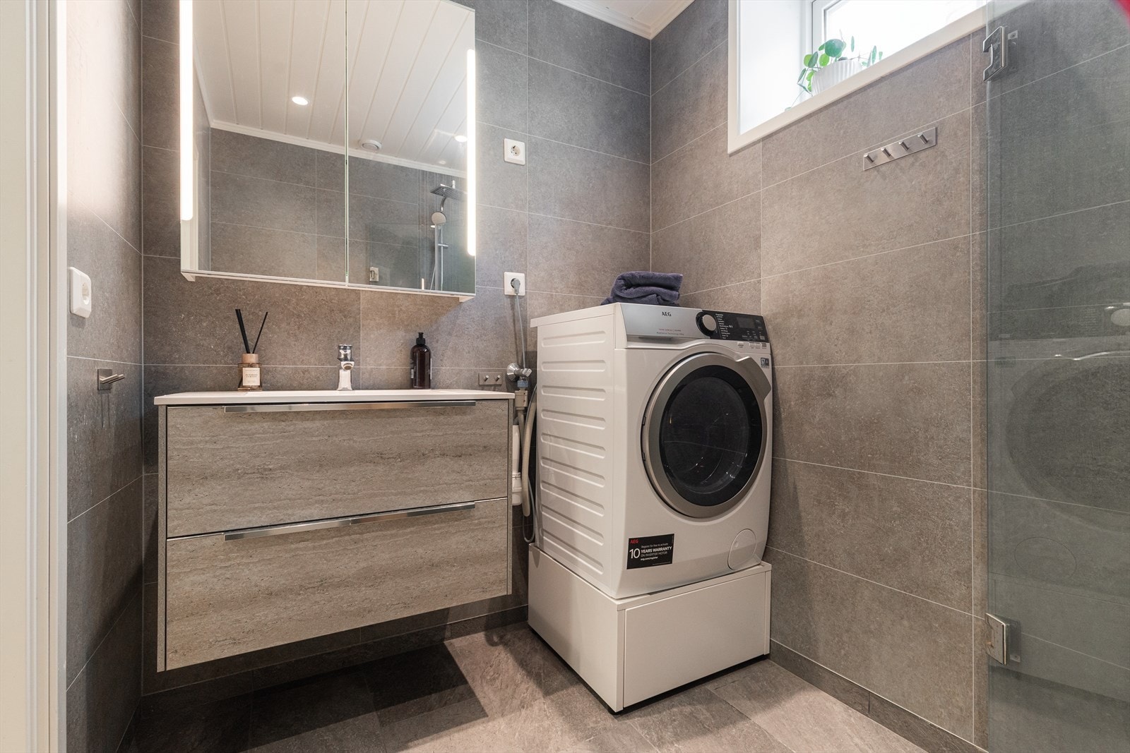 Badet er stilfullt innredet med speil med integrert belysning, nedfelt servant med tilhørende skuffer, klosett, praktisk dusjnisje og opplegg for vaskemaskin. Galleribilde