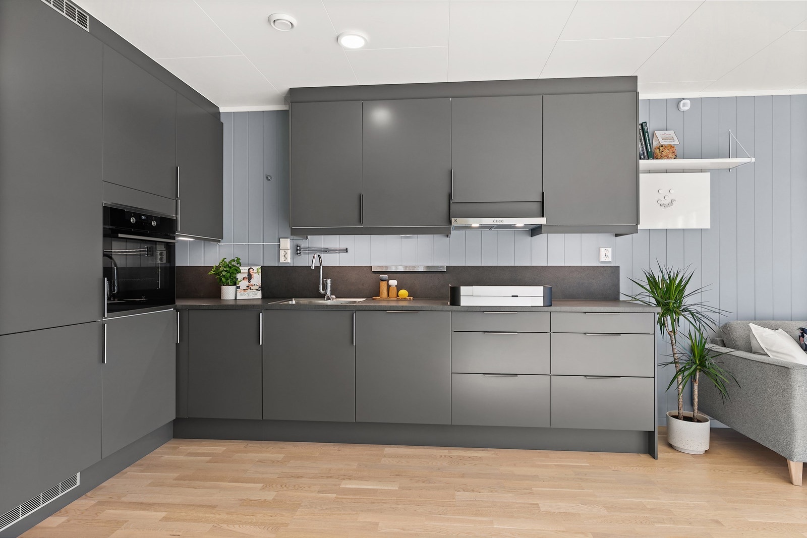 Stilrent og moderne kjøkken i praktisk L-form, med glatte mørkegrå fronter og en matchende veggplate som strekker seg elegant opp fra benkeplaten. Galleribilde