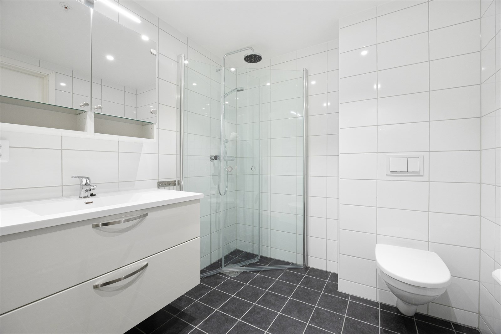 Badet har klassiske hvite fliser på vegger og grå fliser på gulvet. Vegghengt toalett og vannbåren gulvvarme. Galleribilde
