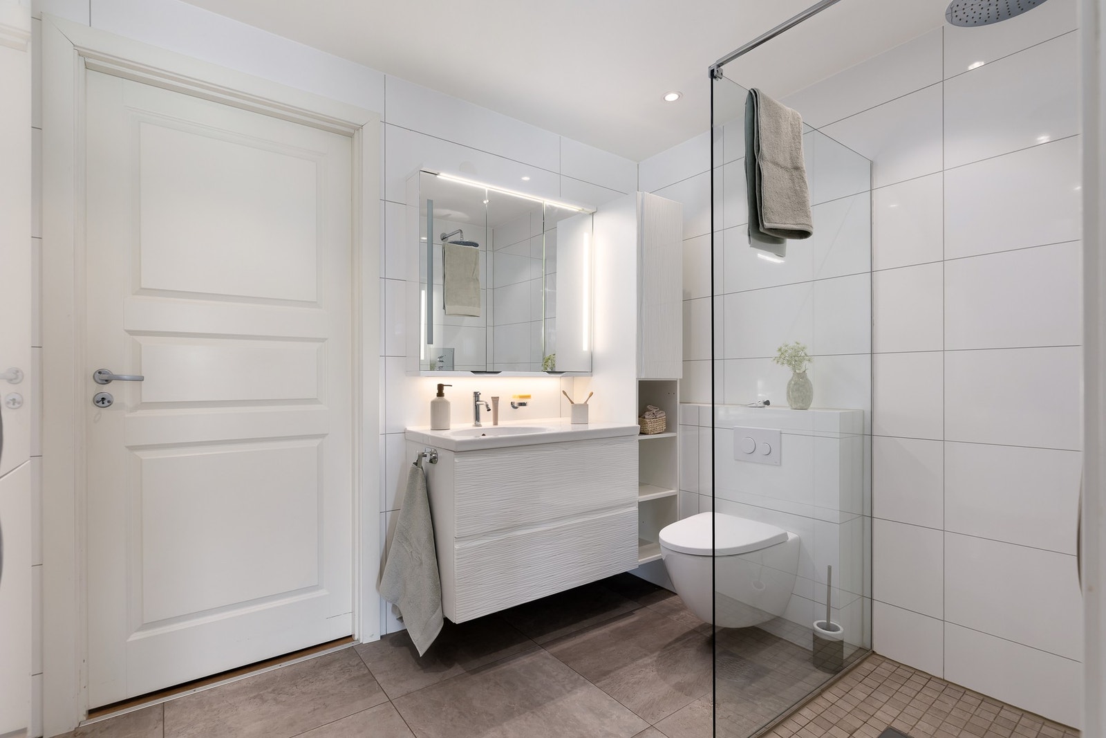 Badet er innredet med vegghengt toalett, servant med underskap, speilskap med overlys, veggskap og åpen dusjløsning med glassvegg. Galleribilde