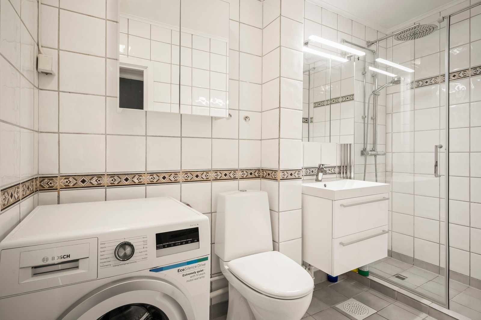 Badet har fliser på både gulv og vegger med varmekabler i gulvet. Baderomsinnredningen består av gulvstående toalett, dusjhjørne og enkel servant. Galleribilde