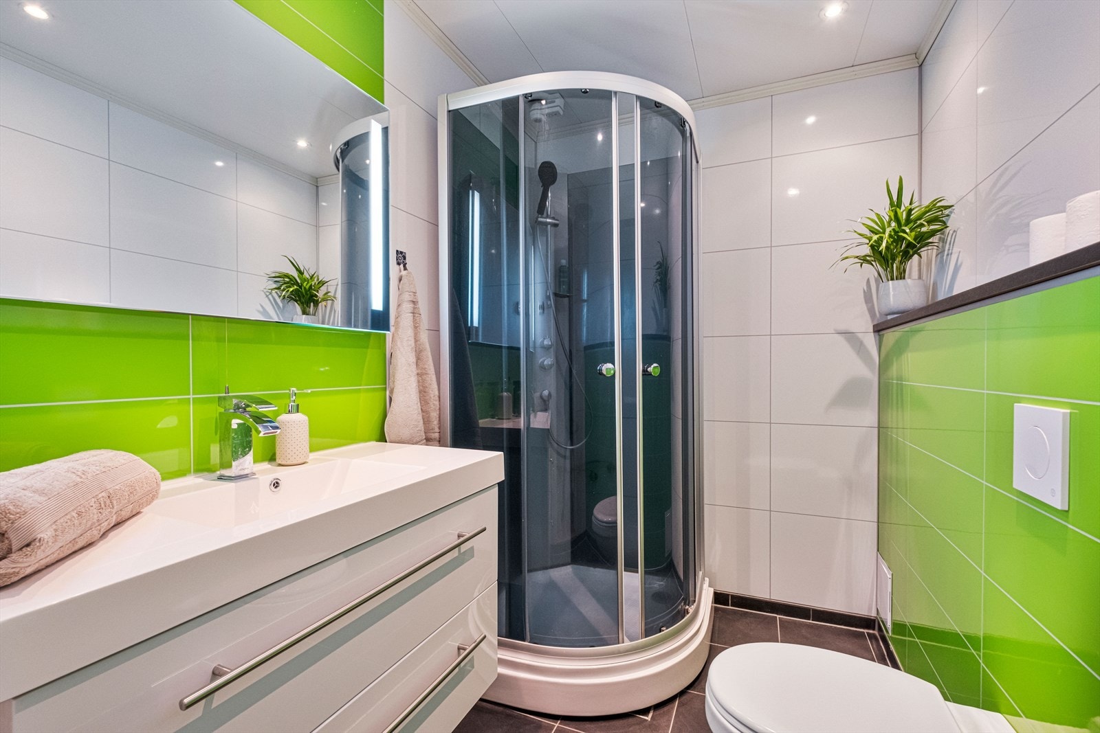 Bad med freshe farger og fliser på gulv med varmekabler. Badet er utstyrt med servant, speil med lys, veggmontert toalett og dusjkabinet. Galleribilde