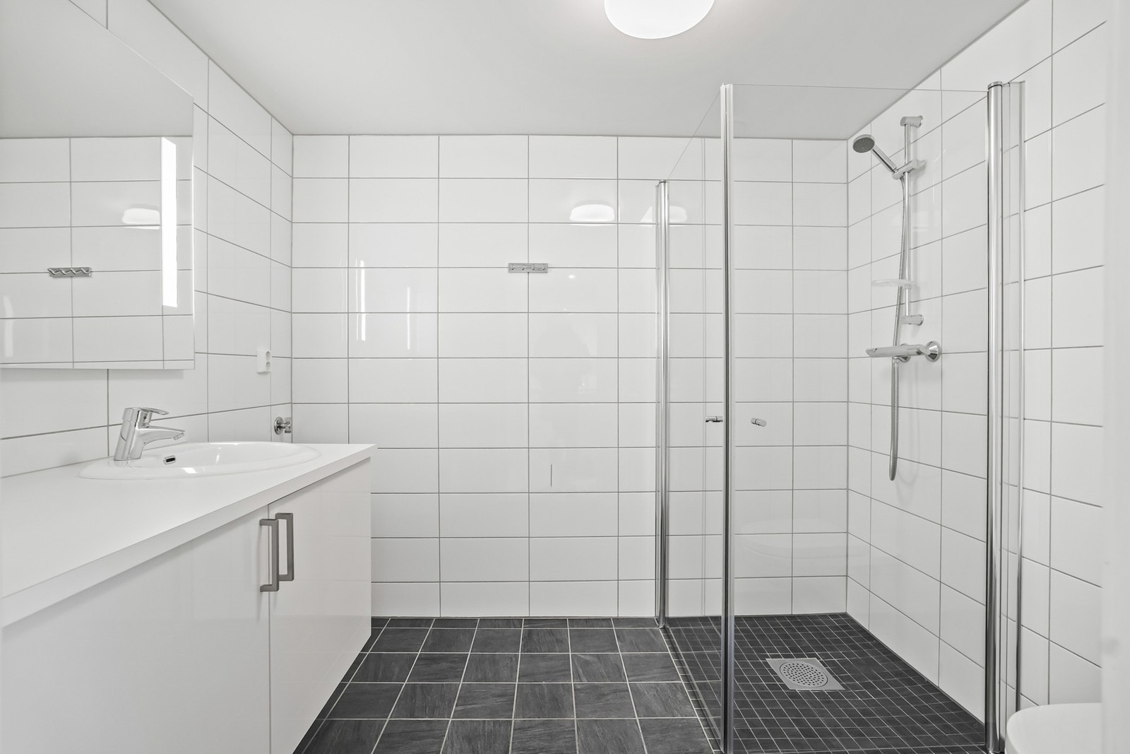 Badet er flislagt og innredet med servantskap, speil, vegghengt toalett og dusjhjørne med glassvegger. Opplegg for vaskemaskin. Galleribilde