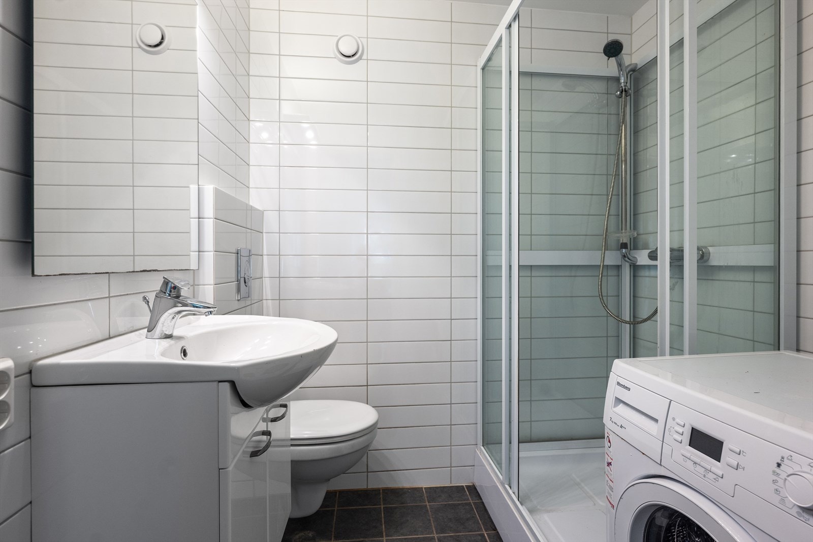 Badet har grå fliser på gulv med gulvvarme og hvite fliser på veggene, kombinert med malt himling. Rommet er utstyrt med dusjkabinett, vegghengt toalett med innebygget sisterne, servantskap med hvite fronter og keramisk servant med ettgreps armatur i krom Galleribilde
