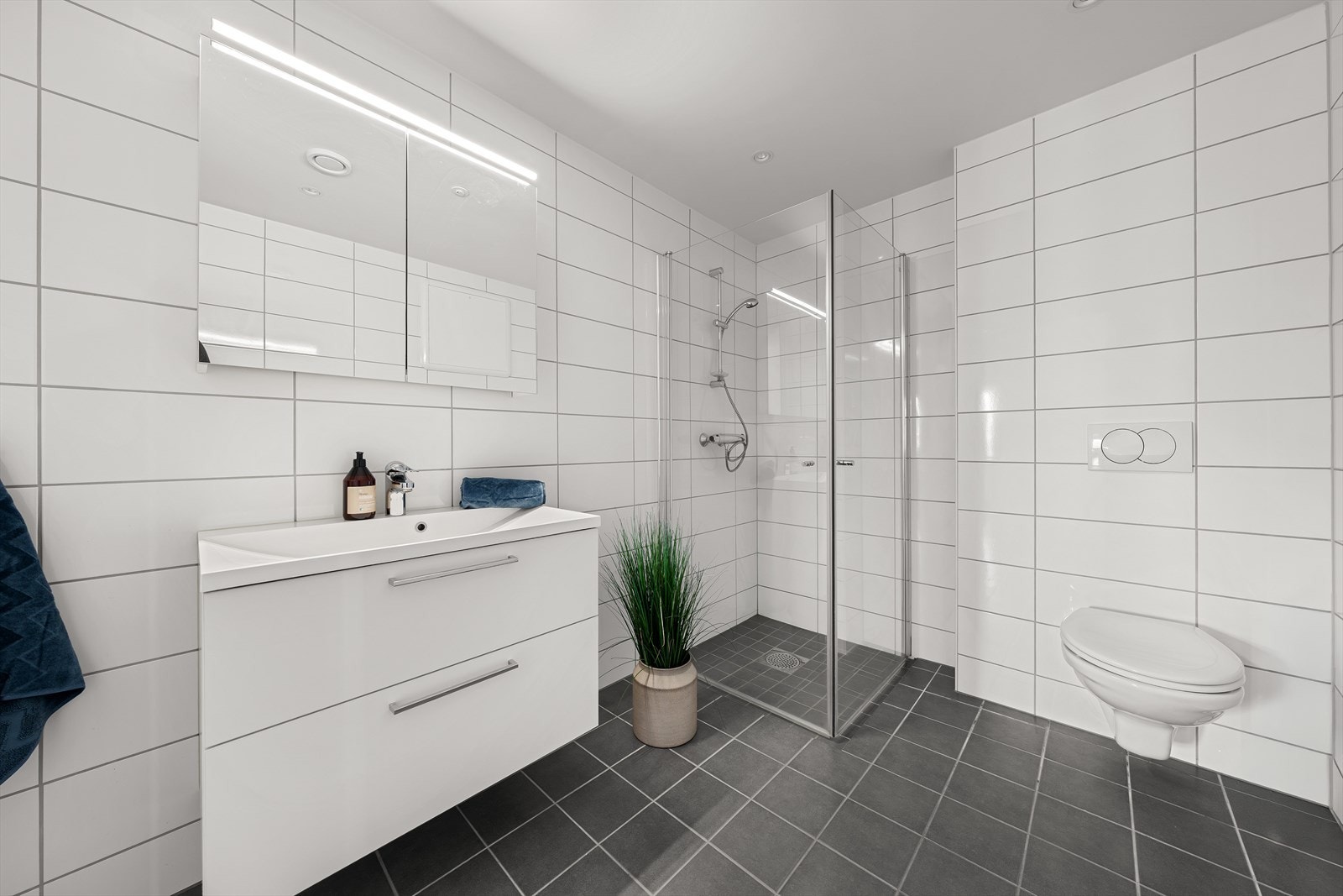 Tiltalende, helfliset baderom med gulvvarme. Badet er utstyrt med heldekkende servant over skuffeinnredning, speil med overliggende belysning, vegghengt toalett og dusjhjørne med innfellbare glassvegger Galleribilde