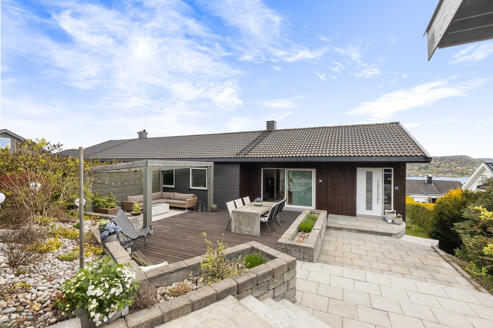 Uteplassen er rammet inn av forstøtningsmurer i betongstein og har integrerte blomsterkasser. Fra stuen er det skyvedør ut til den romslige terrassen. En gjennomtenkt uteplass med ulike soner for sosialt samvær! Galleribilde