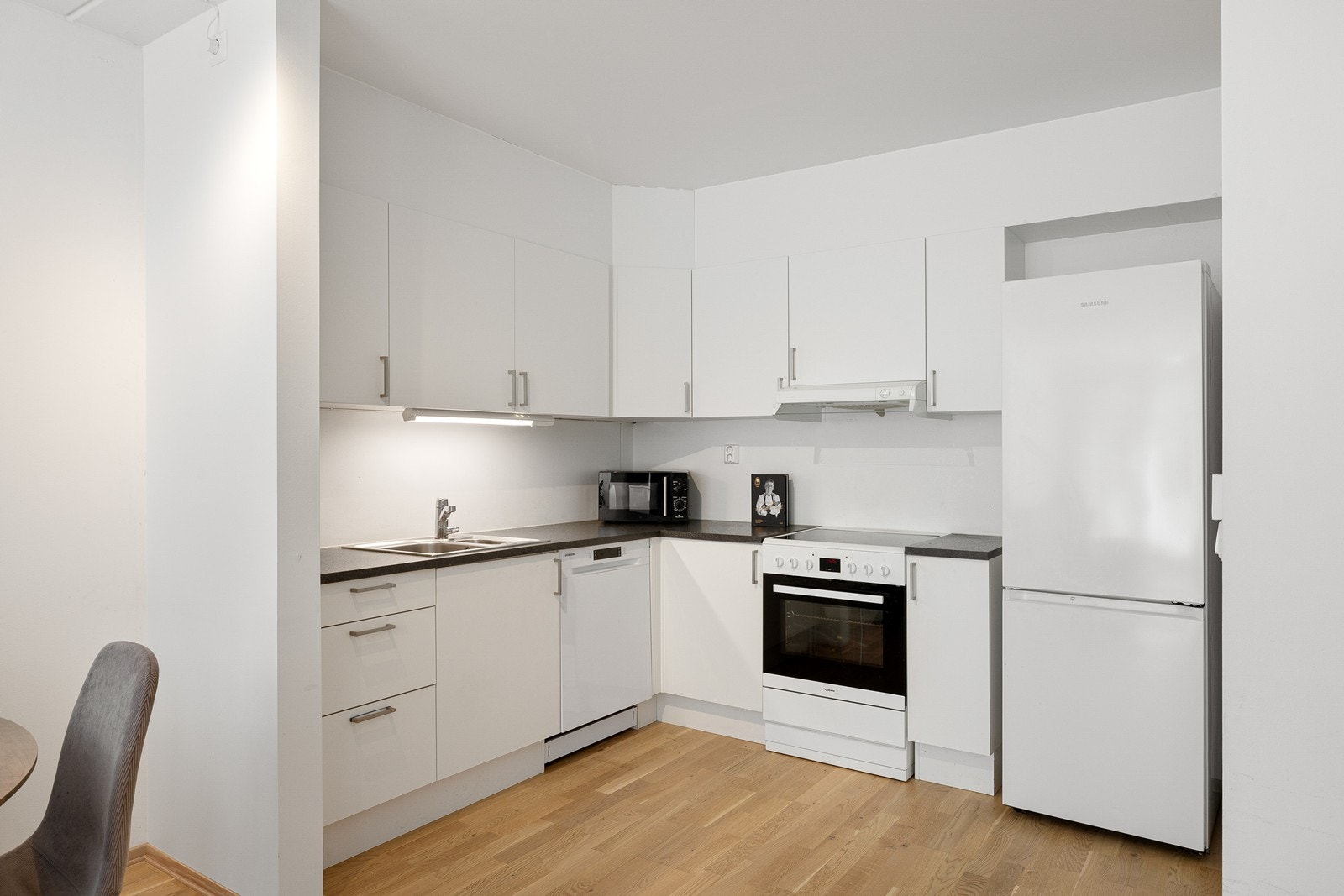 Kjøkkeninnredningen har hvite, glatte fronter som gir et moderne uttrykk, kombinert med en mørk laminert benkeplate med nedfelt oppvaskkum og 1-greps blandebatteri. Galleribilde