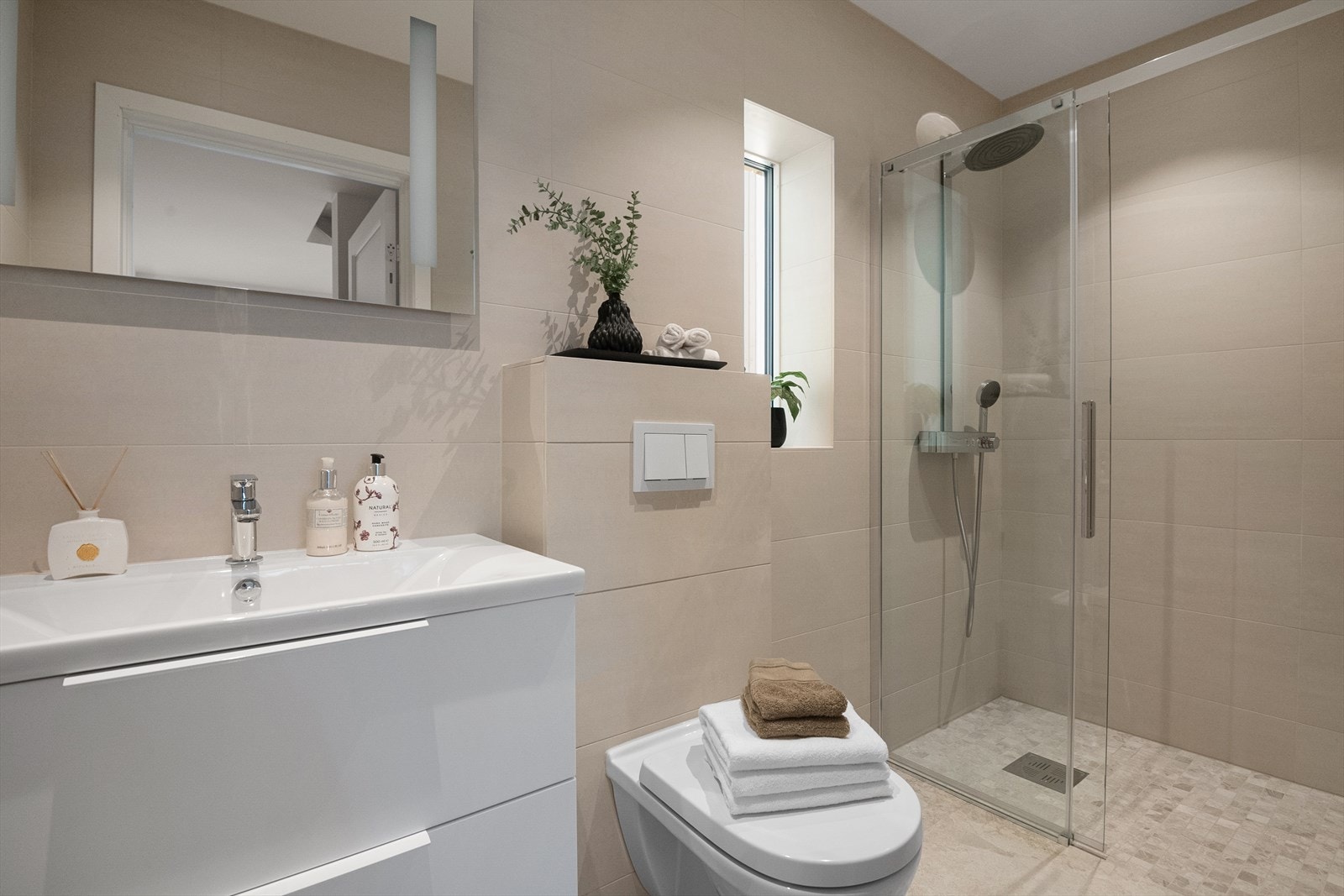 Badet i 1. etasje fremstår moderne og tidløst, med helflisede flater i rolige toner. Dusjhjørne med glass, vegghengt toalett og servantskap gir et stramt og elegant uttrykk. Varmekabler i gulvet og balansert ventilasjon gir høy komfort. Galleribilde