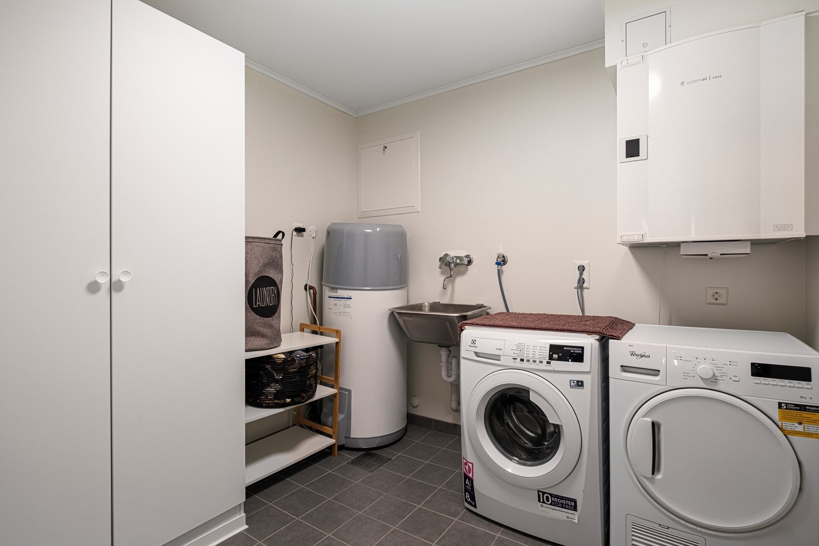 Praktisk vaskerom med fliser på gulv og varmekabler. Vaskerommet er utstyrt med opplegg for vaskemaskin, varmtvannsbereder, sikringsskap, rørskap, utslagsvask og balansert ventilasjon. Galleribilde