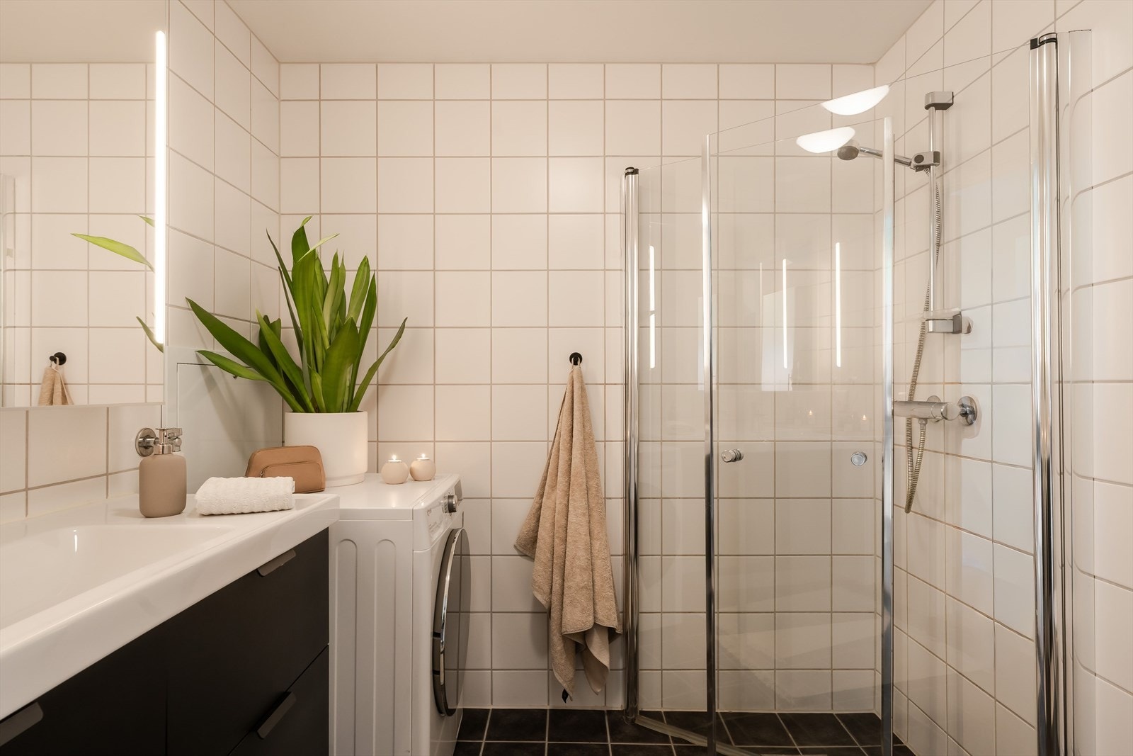 Badet er godt utnyttet, med dusjdører som kan felles innover, servant med underskap og opplegg for vaskemaskin. Veggene er flislagt med hvite fliser, mens gulvet har mørke fliser, noe som gir et stilrent og moderne uttrykk. Galleribilde