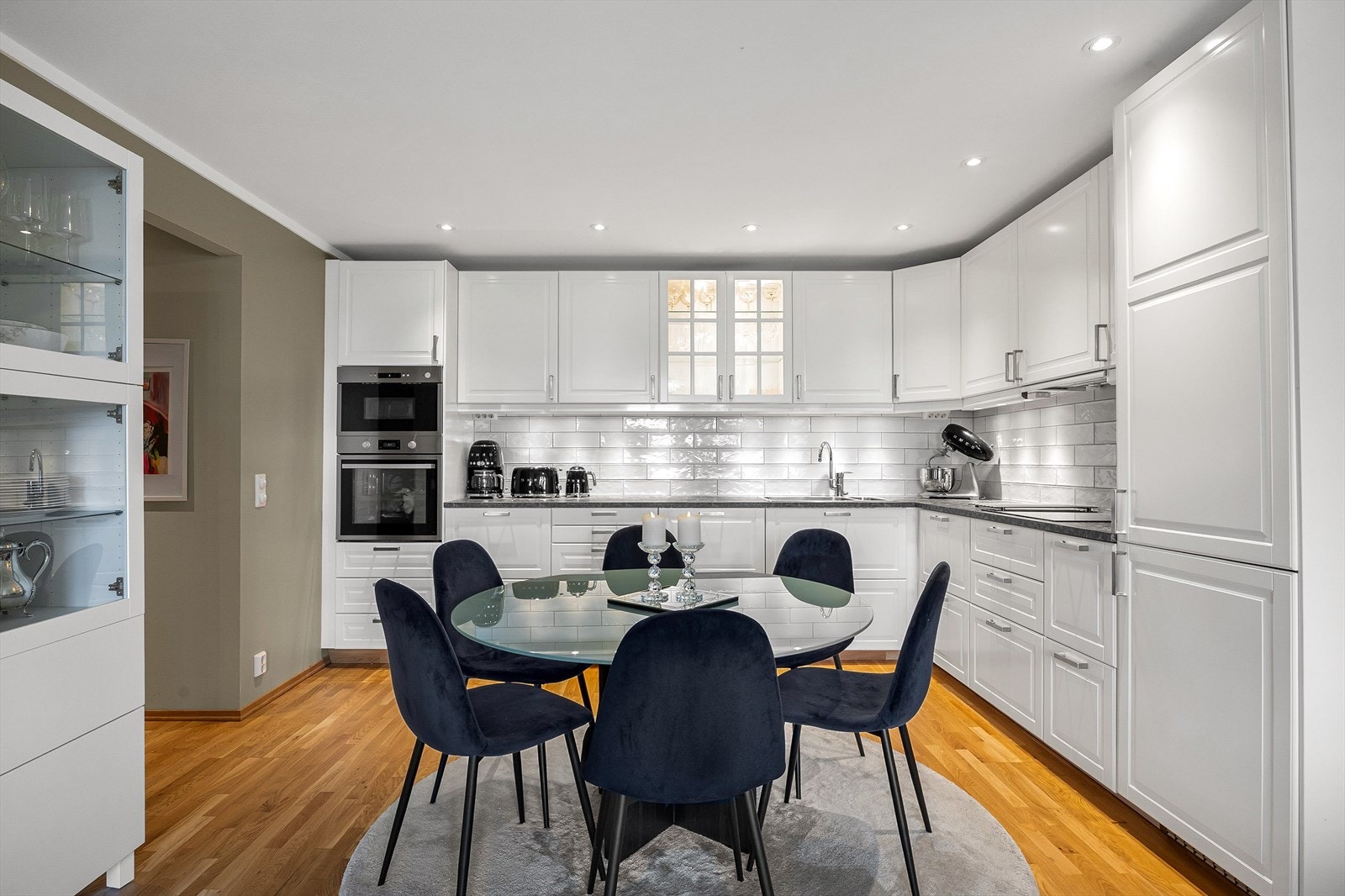 Kjøkkeninnredningen har hvite, profilerte fronter og laminert benkeplate. Ett overskap har glassfronter og integrert belysning, og det er montert downlights i himling over kjøkkeninnredningen. Galleribilde