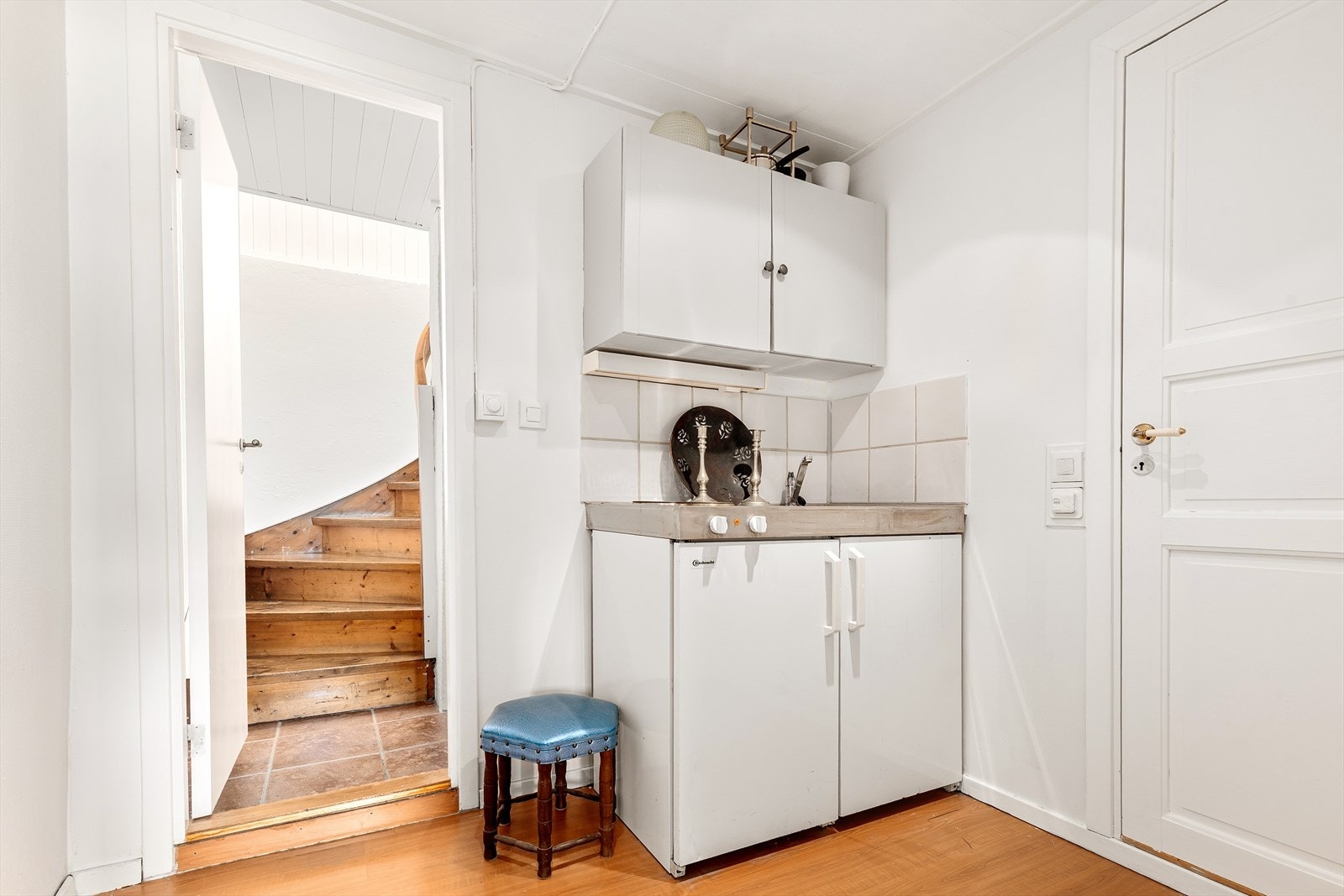 Tekjøkkenet i underetasjen har eldre kjøkkeninnredning med slette, hvite fronter. Det er en heldekkende oppvaskkum i rustfritt stål med avrenningsplate og ettgreps kjøkkenkran. Videre er det installert stekeplate og et integrert benkekjøleskap. Galleribilde