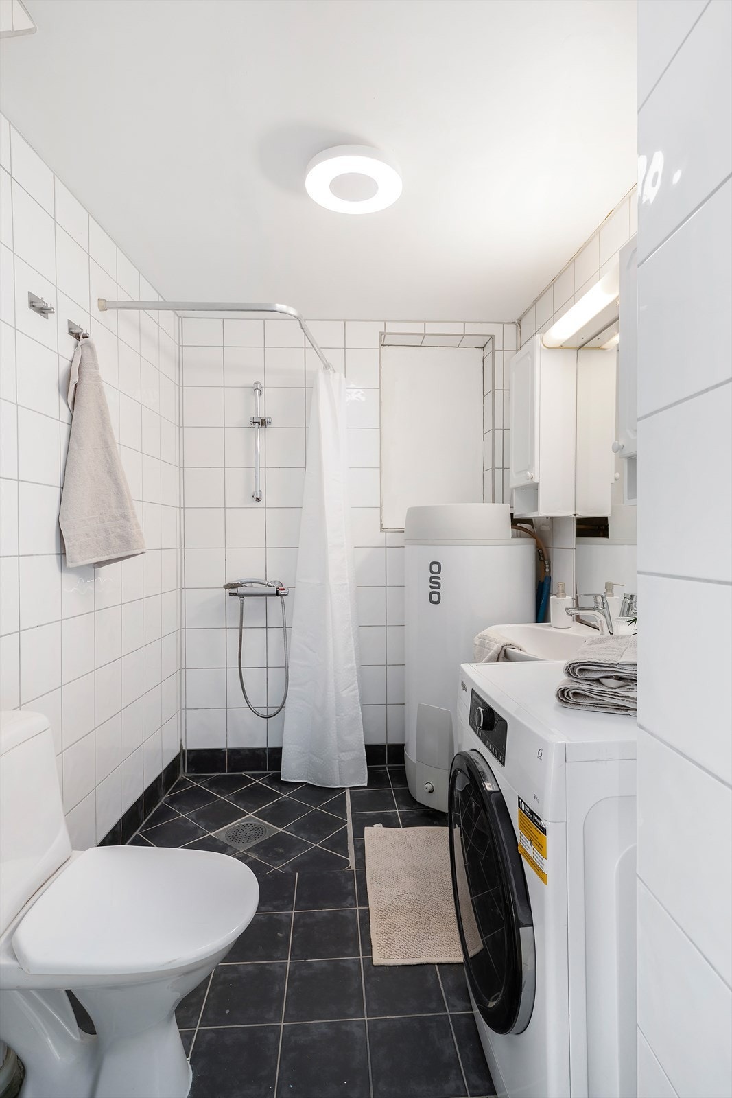 Flislagt bad med servant med tilhørende innredning, toalett, dusjløsning og opplegg for vaskemaskin. Galleribilde