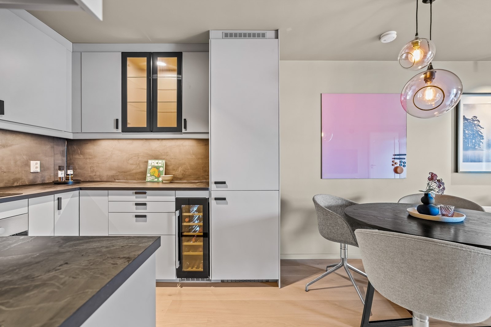 Kjøkkeninnredning med lakkerte fronter, laminat benkeplater, oppvaskkum, ventilator, plater over benk og integrert LED-belysning under overskap. To overskap med fronter i glass gir et elegant preg. Galleribilde