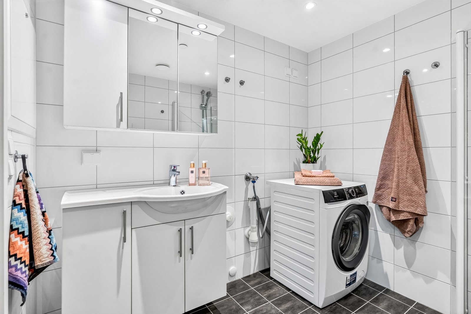 Badet er delikat innredet med mørke gulvfliser og hvite veggfliser. Varme er lagt i gulvet, og badet har downlights, vegghengt toalett og et dusjhjørne med innfellbare glassdører. Galleribilde