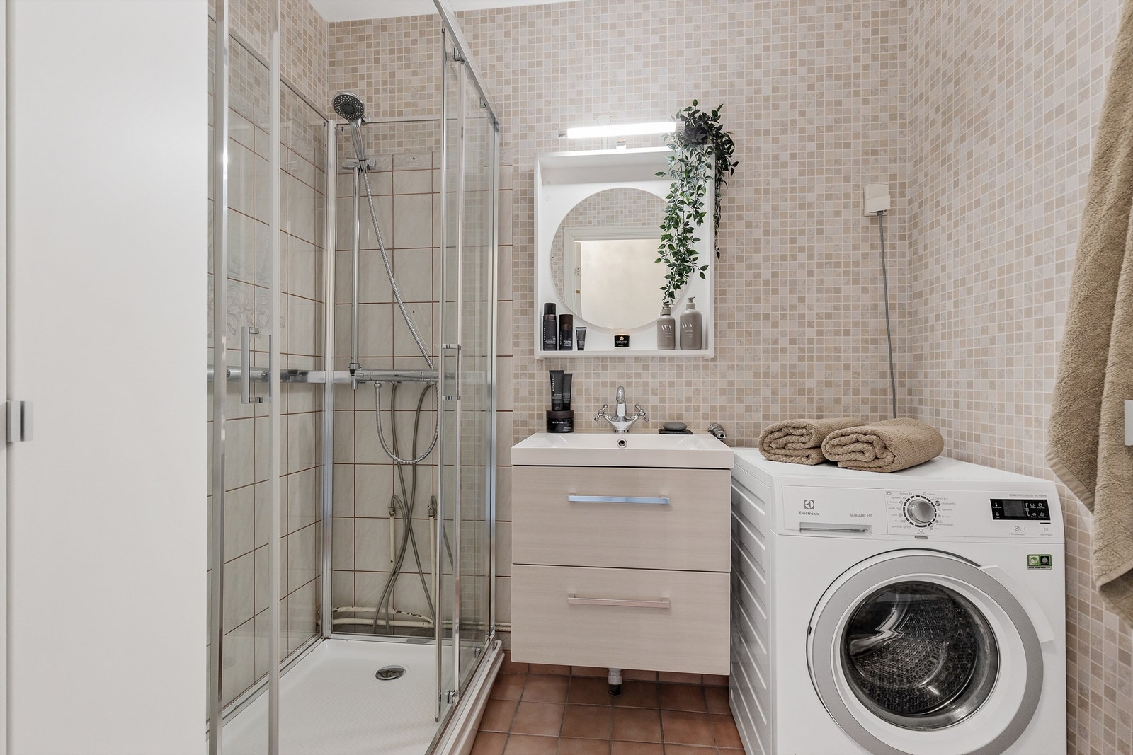 Badet er flislagt på gulv og vegger, utstyrt med dusjkabinett, servantinnredning og opplegg for vaskemaskin. Galleribilde
