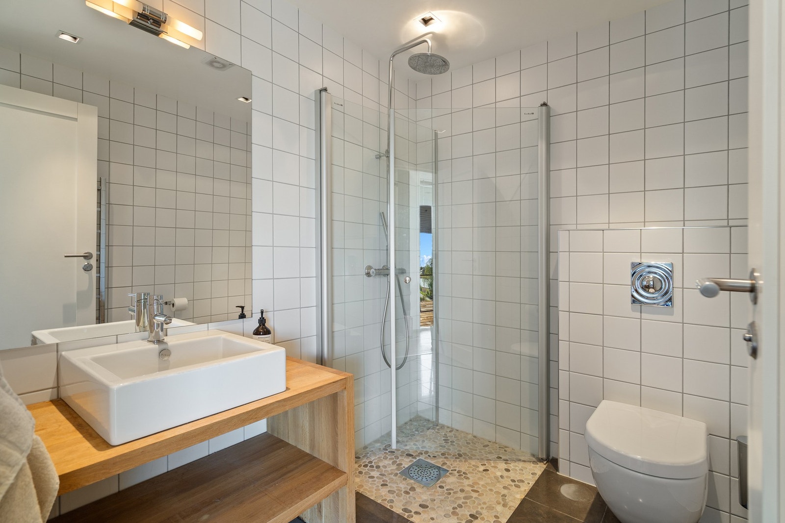 Det andre badet har et lysere og mer minimalistisk preg, med flislagte vegger og gulv, samt dusjsone med glassvegger og detaljer som gir et moderne uttrykk. Servant med benkeplate i tre tilfører varme til rommet og skaper en fin kontrast til de lyse flate Galleribilde