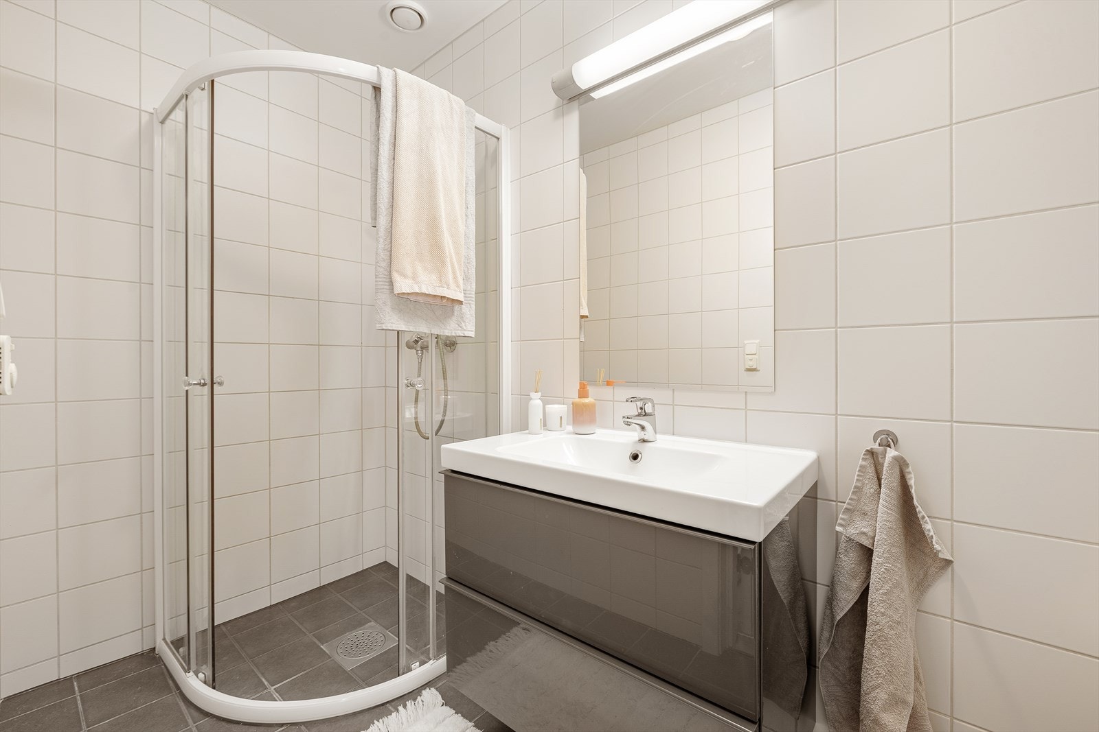 Lyst og flislagt baderom utstyrt med dusjhjørne, servantinnredning, toalett og opplegg for vaskemaskin. Galleribilde