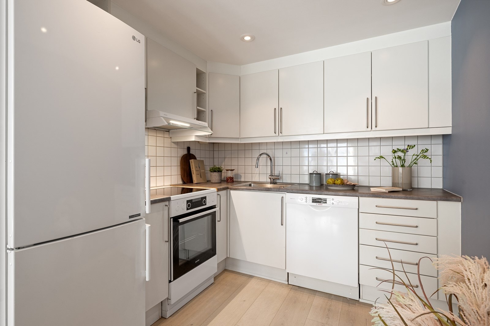 Lys og stilren kjøkkeninnredning med glatte fronter, fliser over benk og laminat benkeplate med nedfelt kum i rustfritt stål. Galleribilde