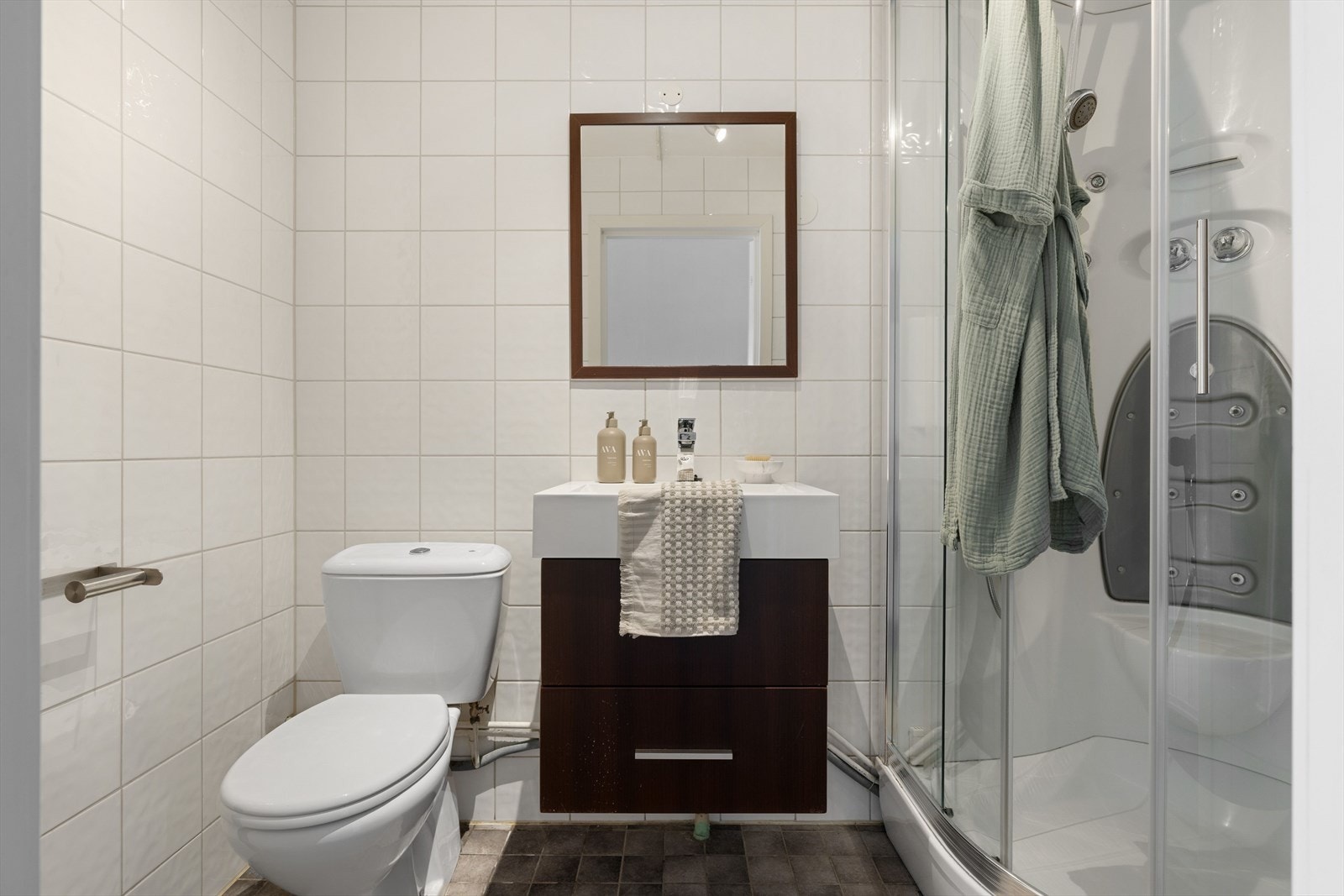Badet har flislagte overflater og er utstyrt med toalett, dusjkabinett og en pen baderomsinnredning med skuffeseksjoner. Galleribilde